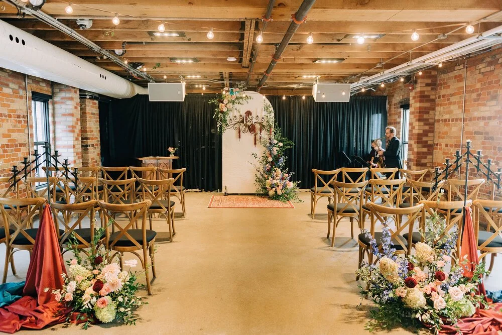 Indoor wedding or event venue with wooden chairs arranged facing an altar decorated with flowers and greenery, black curtains backdrop, brick walls, wooden ceiling with string lights, and two speakers mounted on the ceiling. Two people are near the altar.