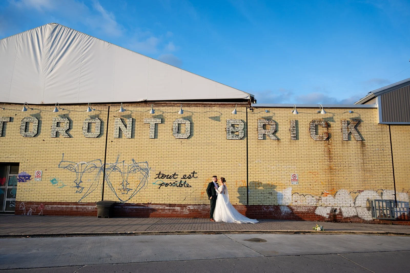 A bride and groom standing on a sidewalk, holding hands, in front of a yellow brick building with graffiti and the words 'TORONTO BRICK' on it. The bride is wearing a white wedding dress, and the groom is wearing a black suit. The sky is partly cloudy with patches of blue.