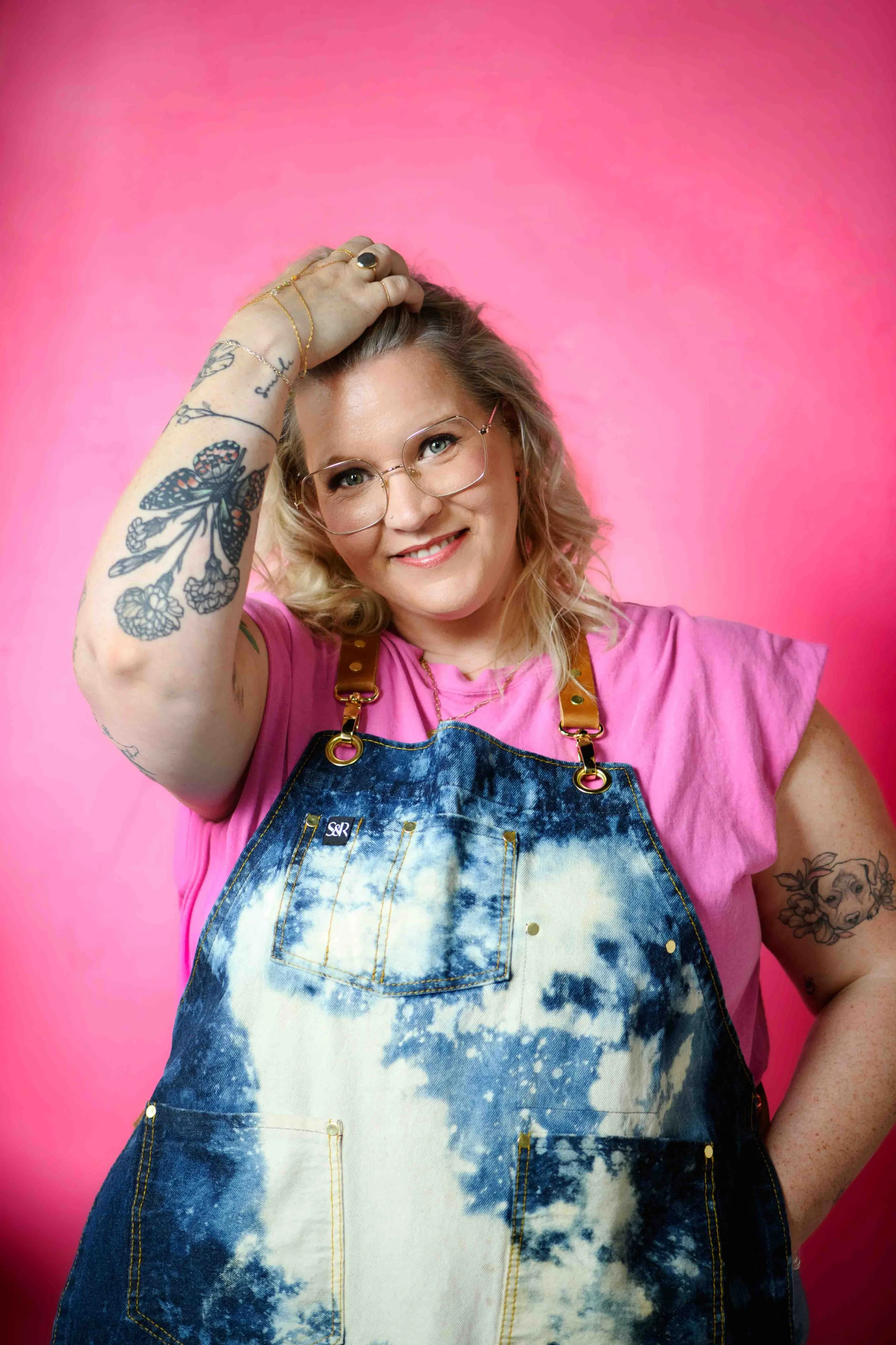 A woman with blonde curly hair, glasses, and tattoos on her arms, wearing a pink t-shirt and tie-dye denim apron, smiling against a pink background.