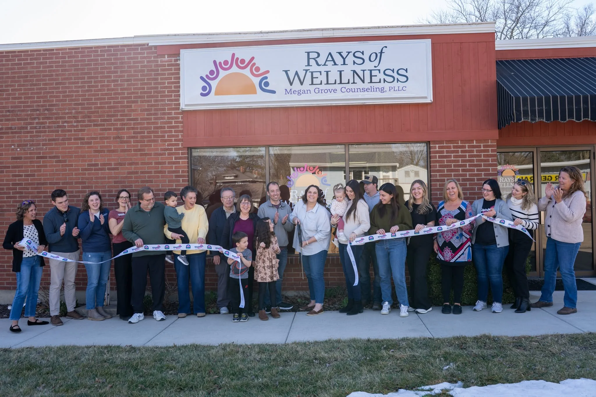 A large group of people holding and cutting a ceremonial ribbon outside the brick office building of Rays of Wellness during a grand opening celebration.