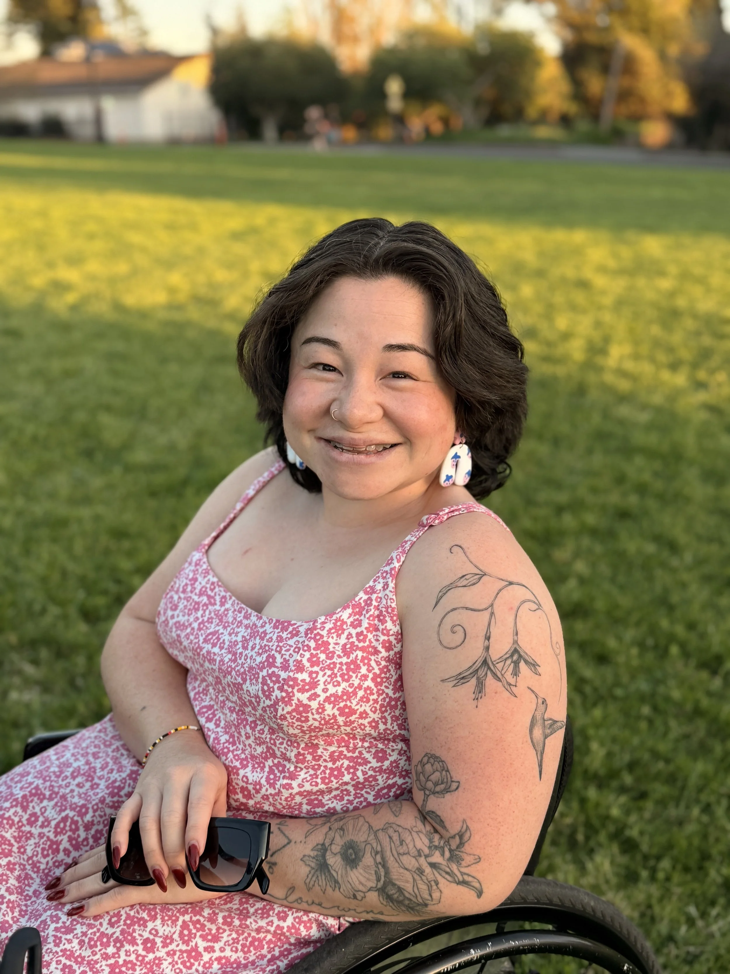 Headshot of Rosemary seated in her manual wheelchair. She is smiling wearing a pink & white dress. She has tattoos on her arm and matching pink earrings.
