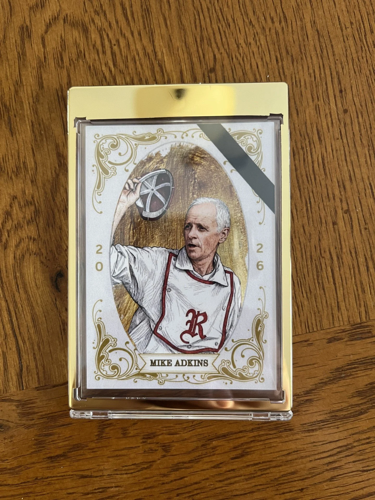 A collectible card featuring Mike Adkins, dressed as a chef, holding a cooking hat. The card has a vintage style with gold decorative borders, the year 2026, and his name at the bottom.