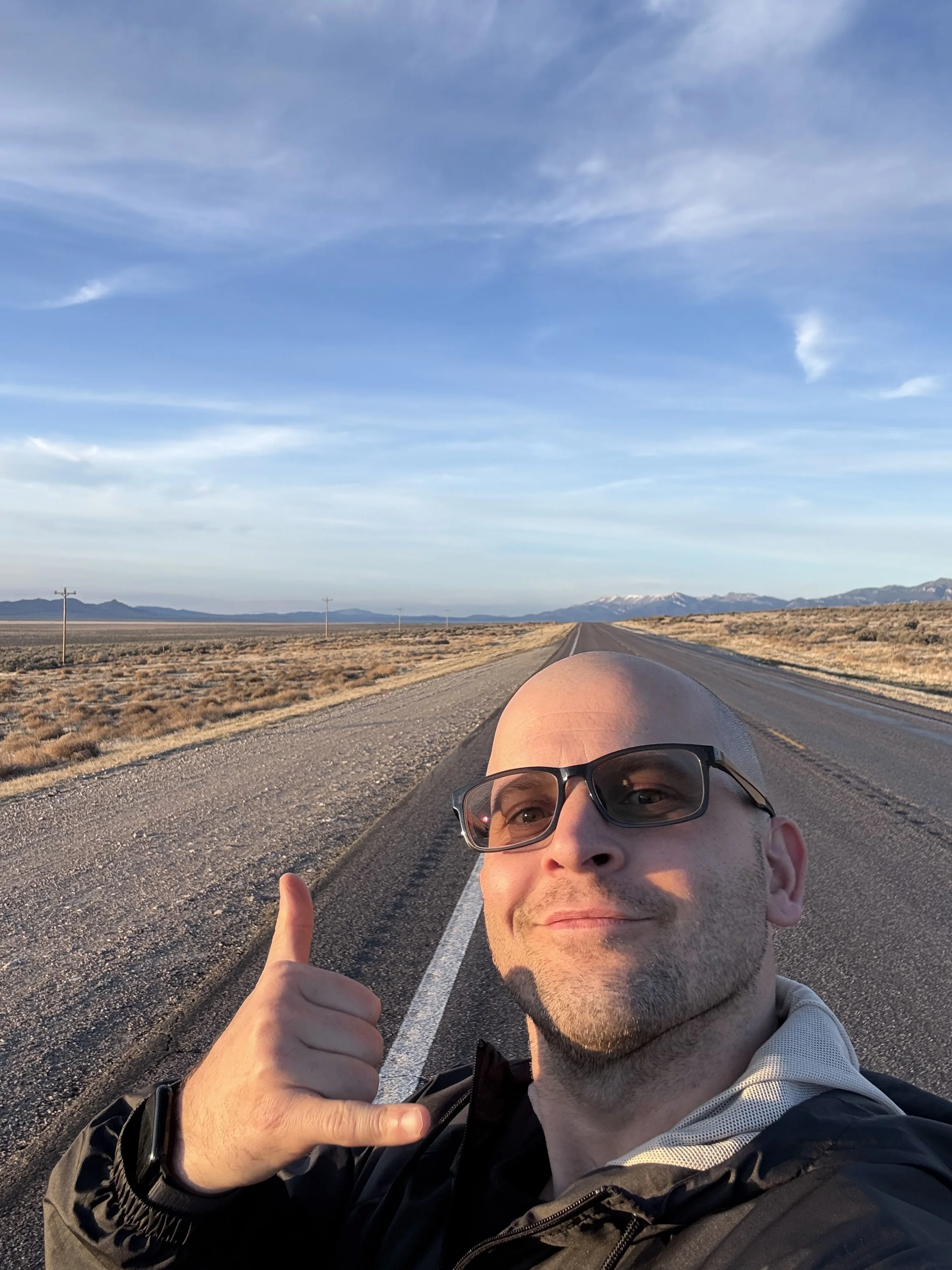A man with glasses taking a selfie on an empty desert road with mountains in the background.