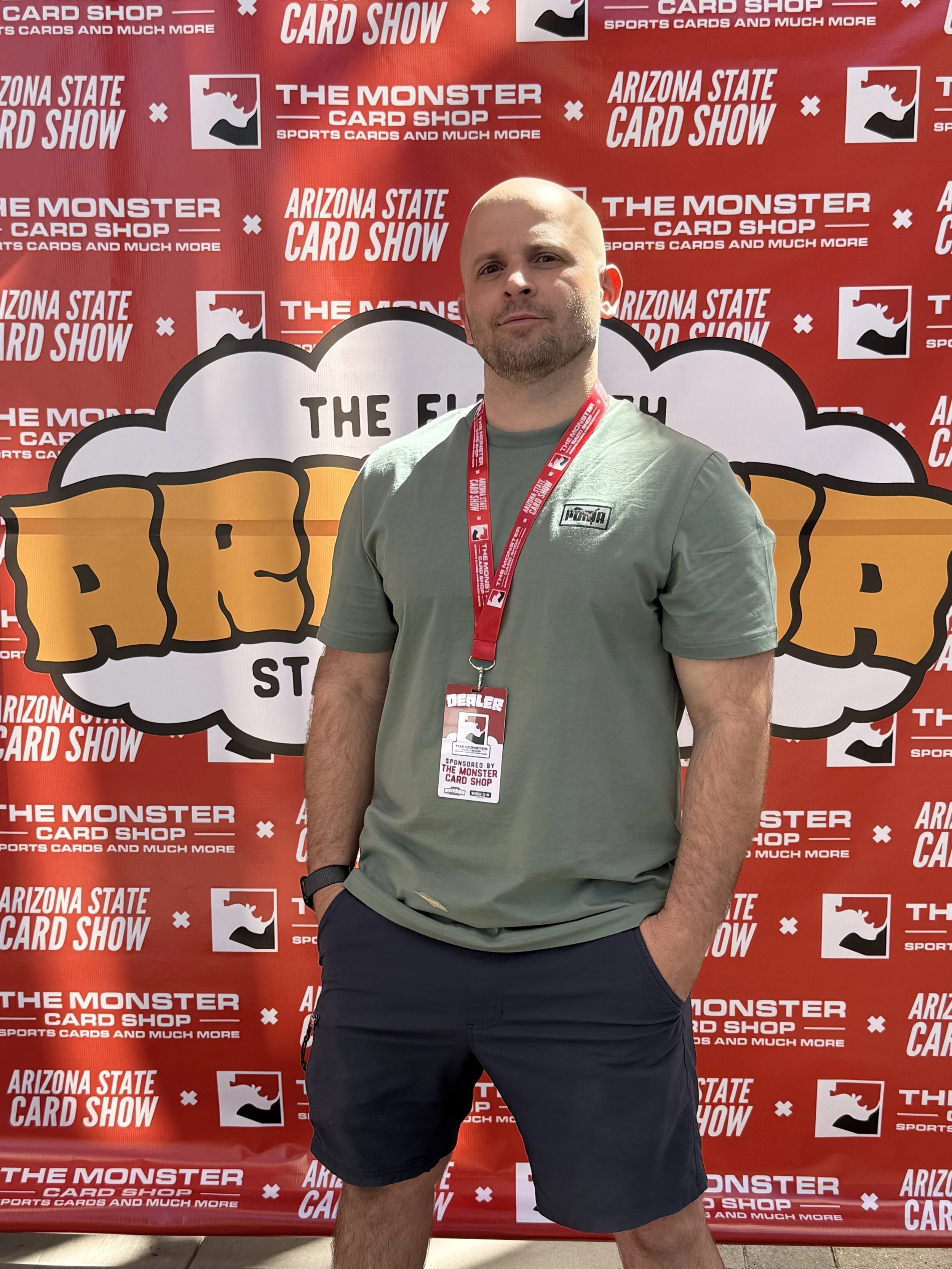 A man stands in front of a red backdrop with logos and text promoting the Arizona State Card Show and The Monster Card Shop. He is wearing a green t-shirt, navy shorts, a red lanyard with a badge, and has his hands in his pockets.