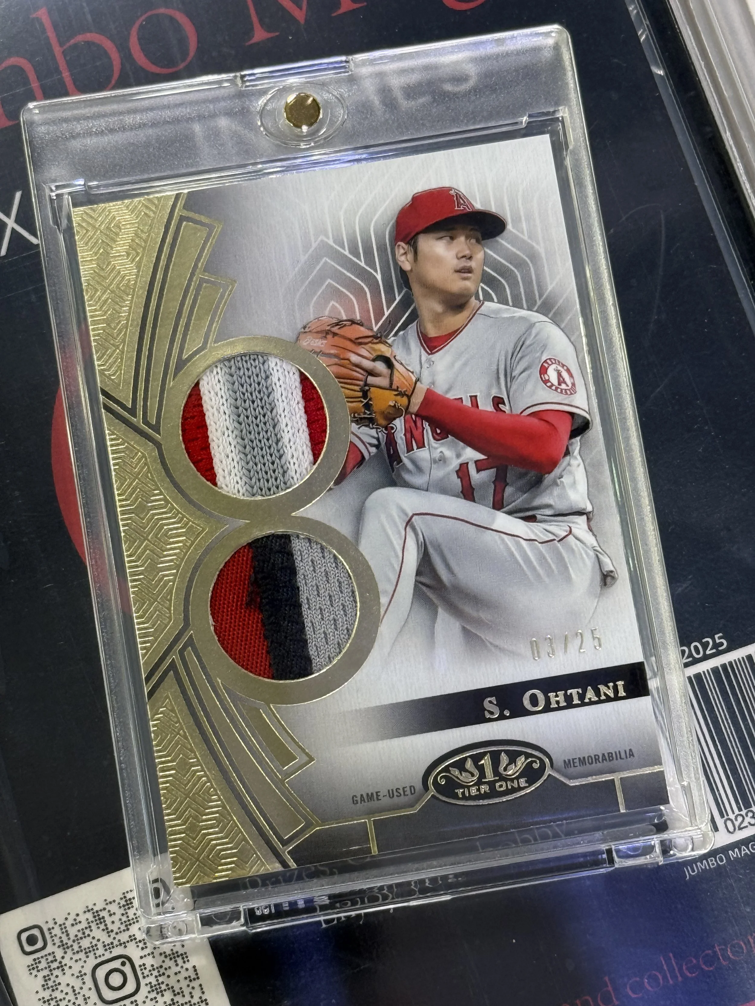 A collectible baseball card featuring a player from the Los Angeles Angels, S. Ohtani, wearing a gray and red uniform and a red cap, holding a baseball glove and kneeling on one knee. The card has patches of fabric from the player's uniform and a dec