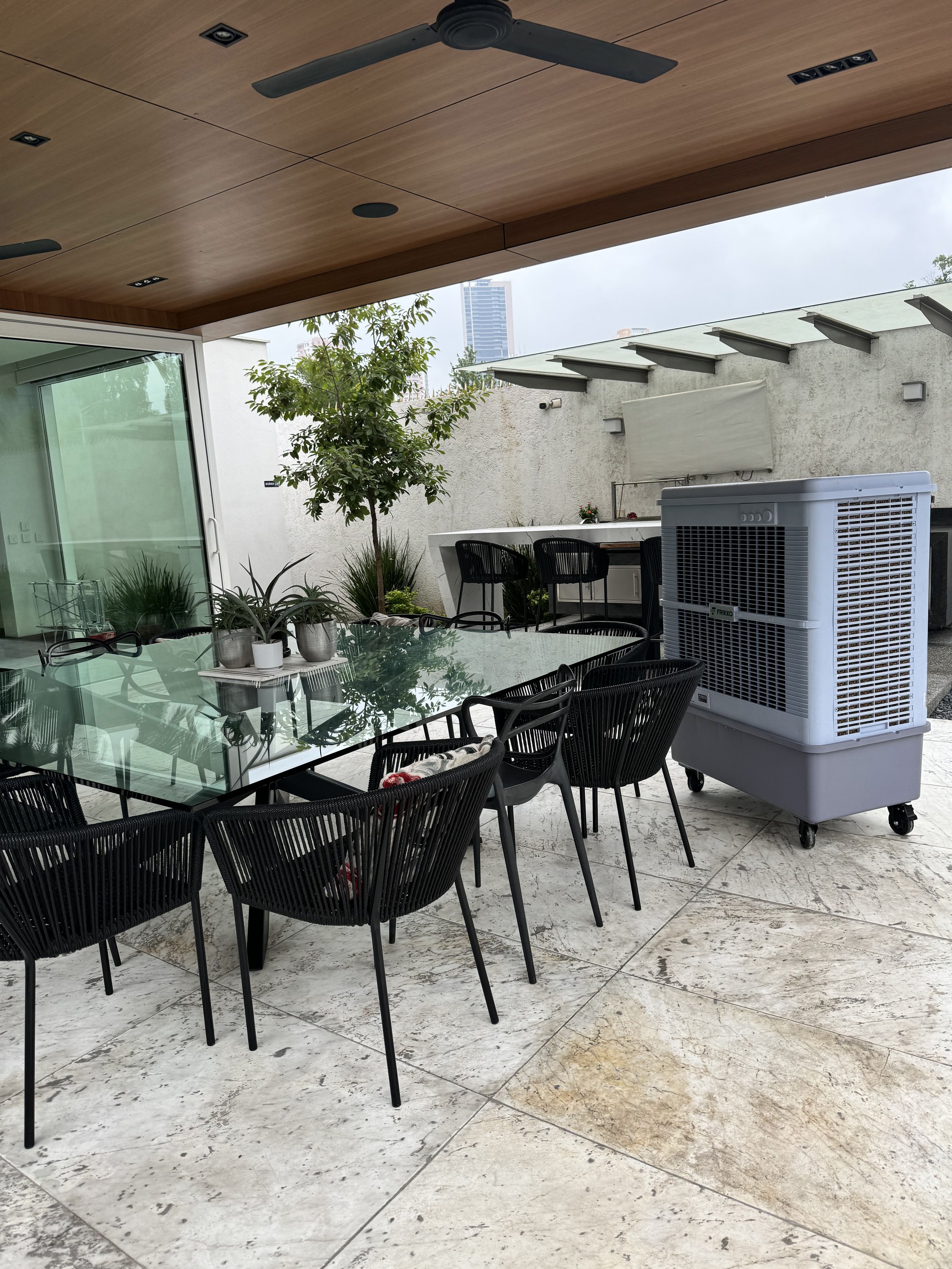 Patio cubierto moderno con mesa de vidrio y sillas negras, ventilador de techo, enfriador de aire portátil y plantas decorativas.