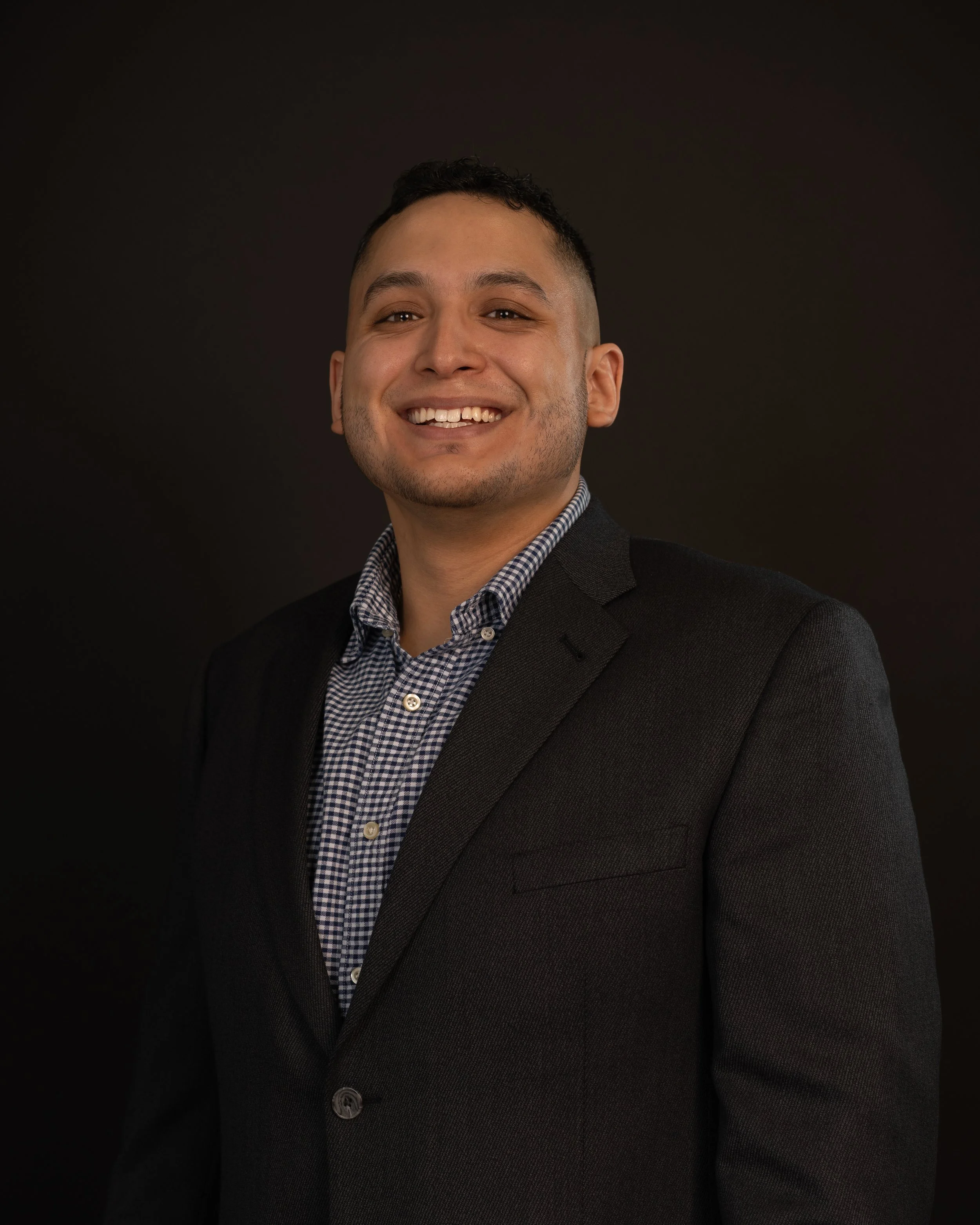 Headshot of a smiling man in a dark suit jacket and checkered shirt against a dark background posing for his professional corporate headshots.