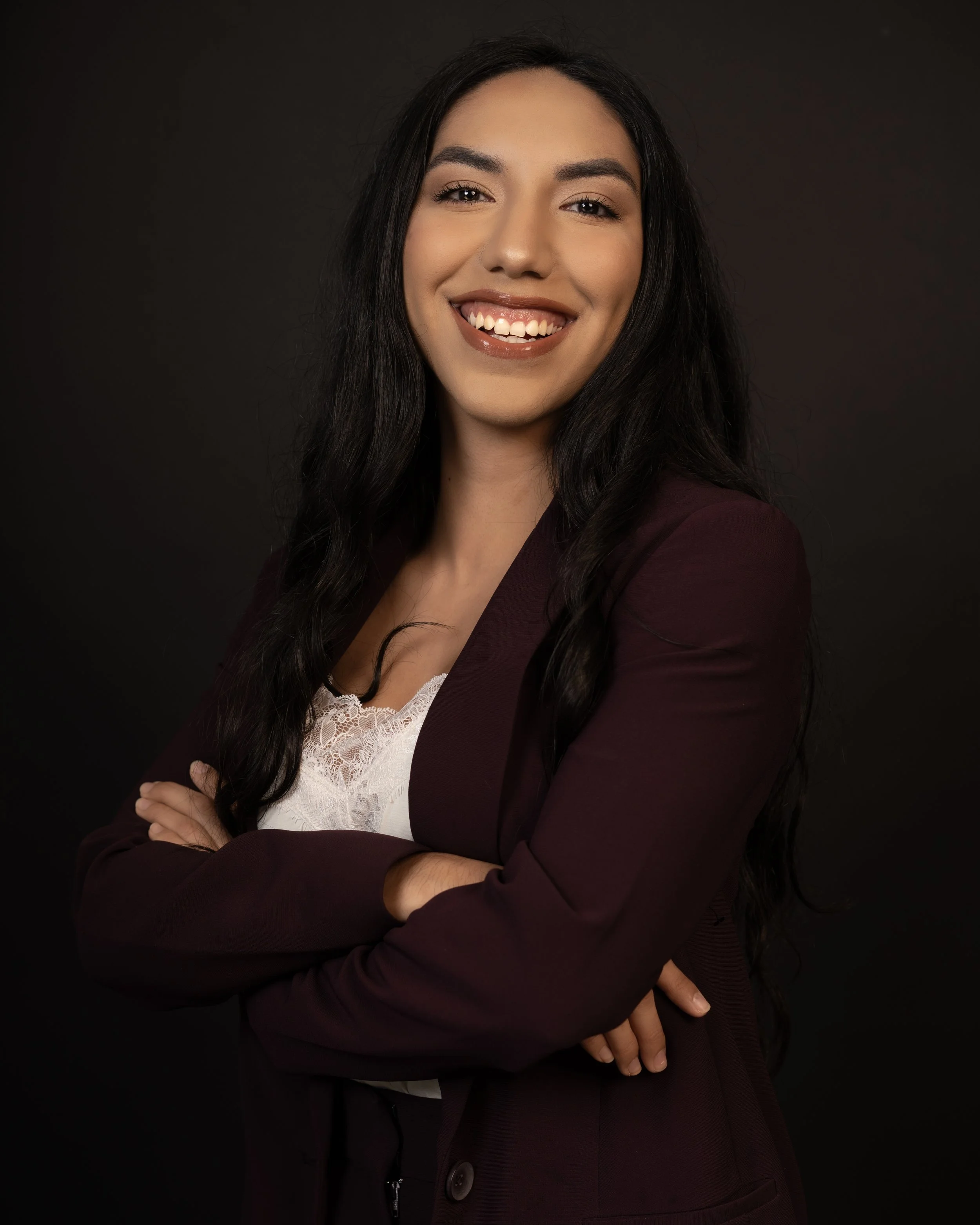 A woman with long dark hair, smiling, with arms crossed, against a dark background posing for her professional headshots taken by Lanza Photography.