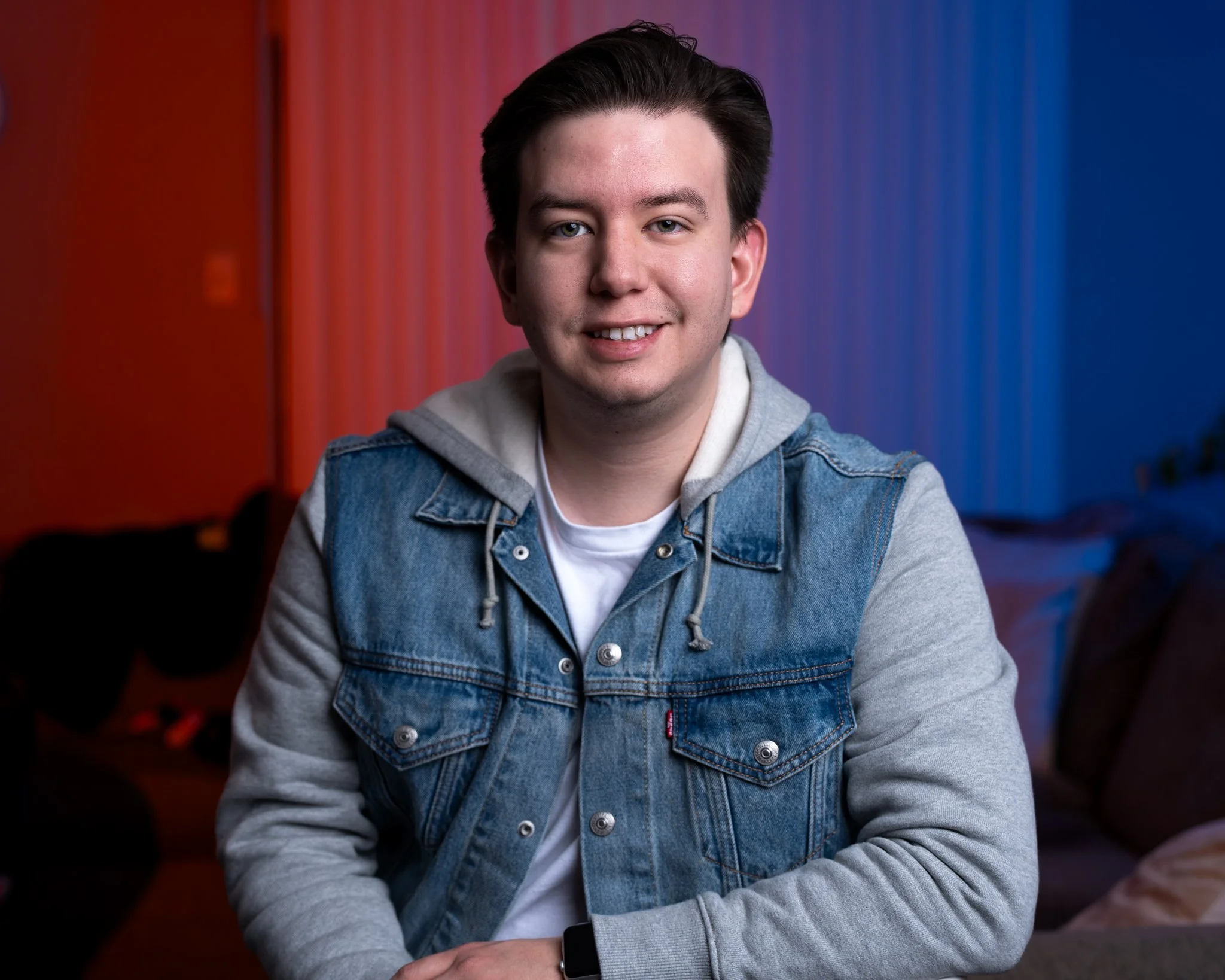 Young professional quinceañera photographer sitting indoors, smiling, wearing a denim and gray hooded sweatshirt, background with red and blue lighting.