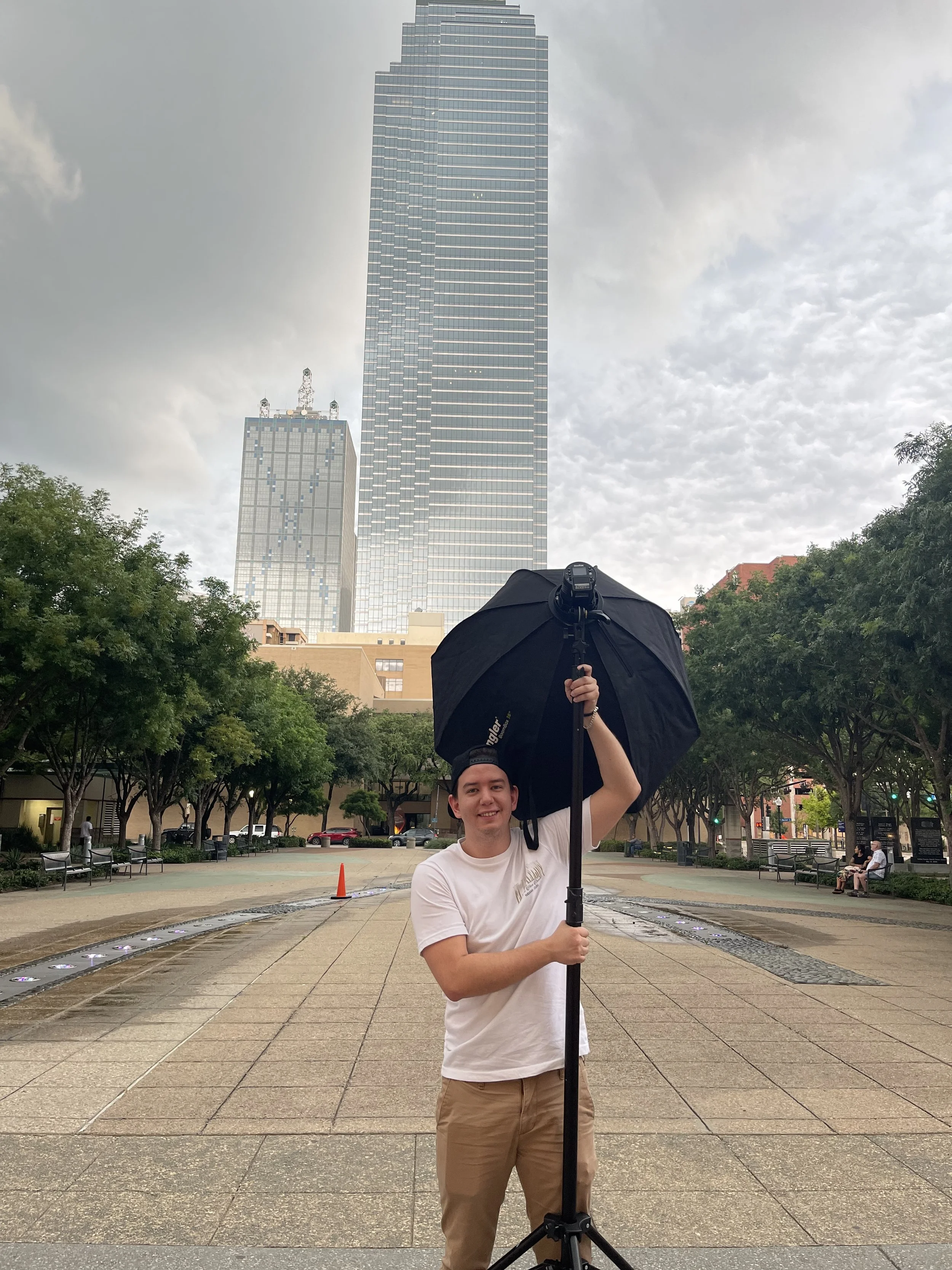 A professional photographer holding a large black umbrella and a camera mounted on a tripod at an outdoor park with tall skyscrapers in the background, and trees lining the scene to take portraits.