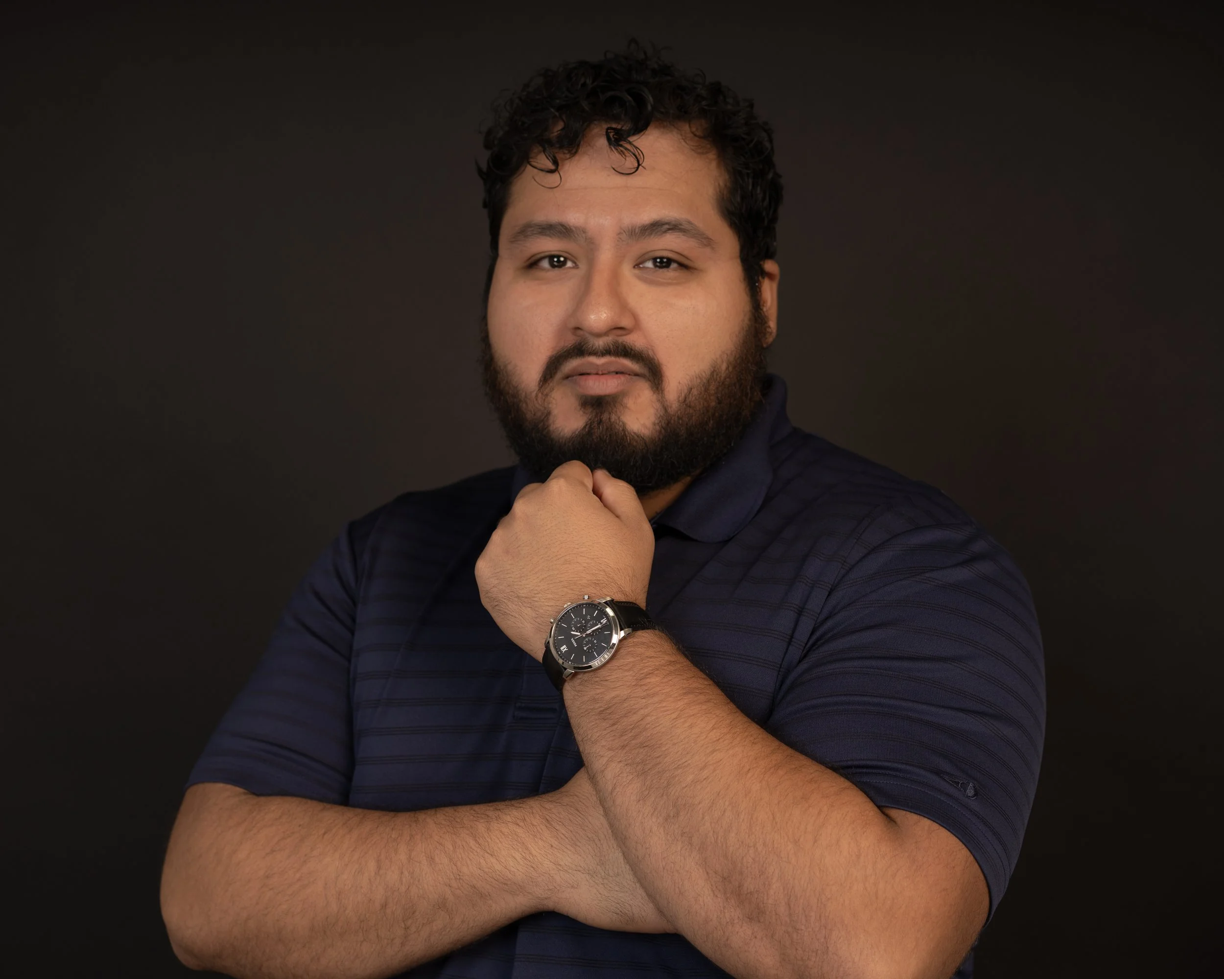 A man with curly hair and a beard wearing a navy blue polo shirt and a black wristwatch, standing against a dark background, with one hand resting on his chin and the other crossed over his chest.