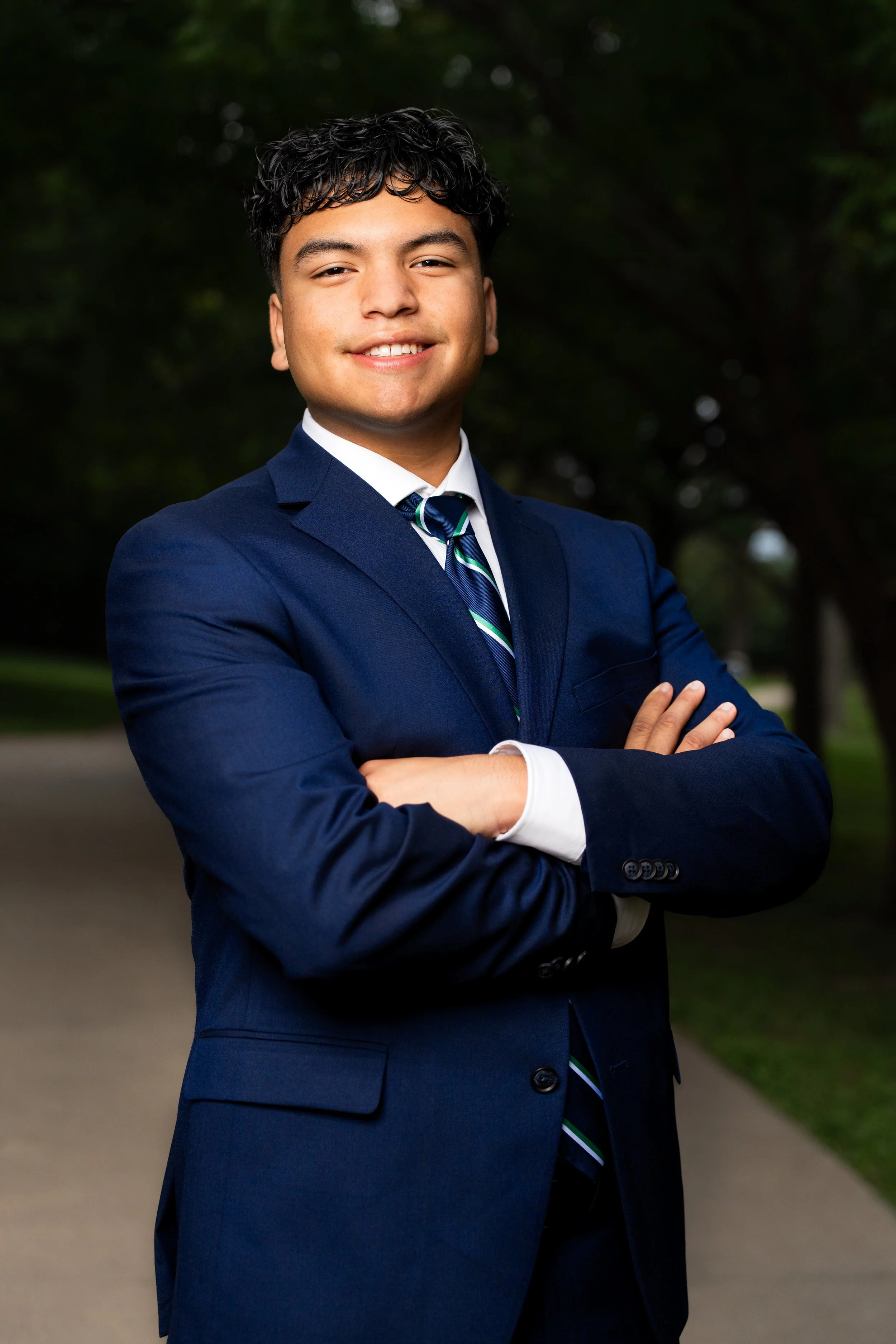 Young man in a navy suit, white shirt, and striped tie, smiling with arms crossed outdoors, trees in the background posing for his graduation senior portraits. 