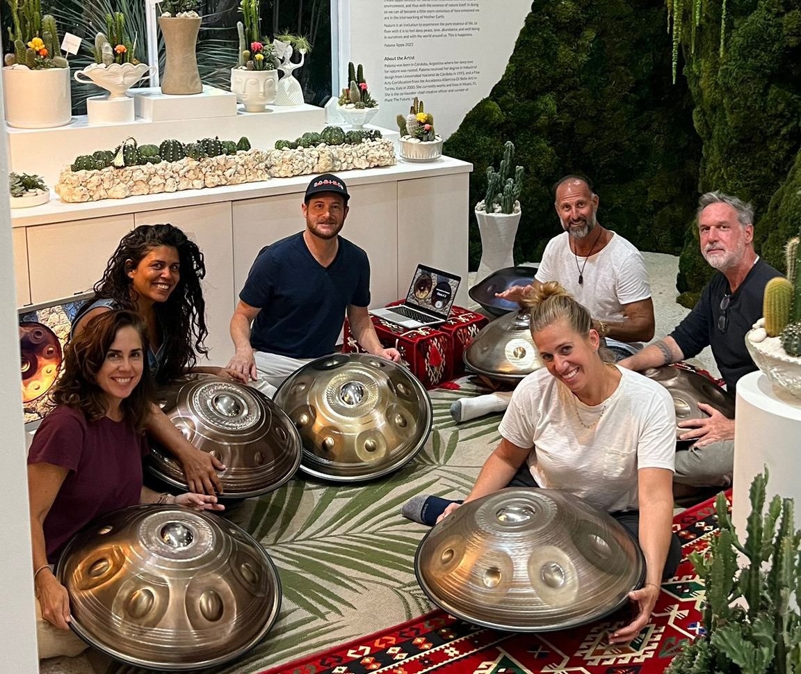 Group of six people sitting on the floor with handpans in a room decorated with plants and cacti.