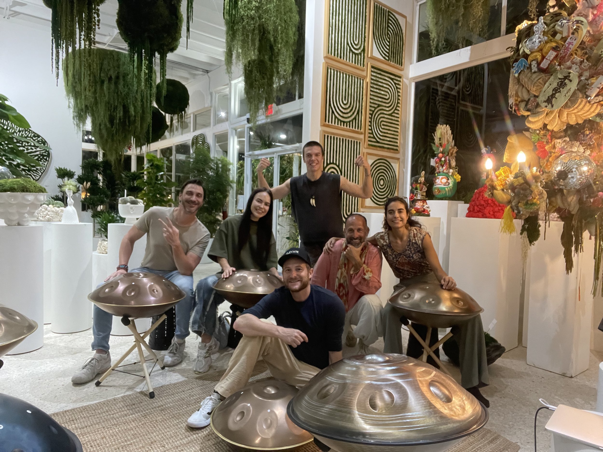Group of six people sitting with hang drums in a plant-filled room, decorated with modern art and sculptures.