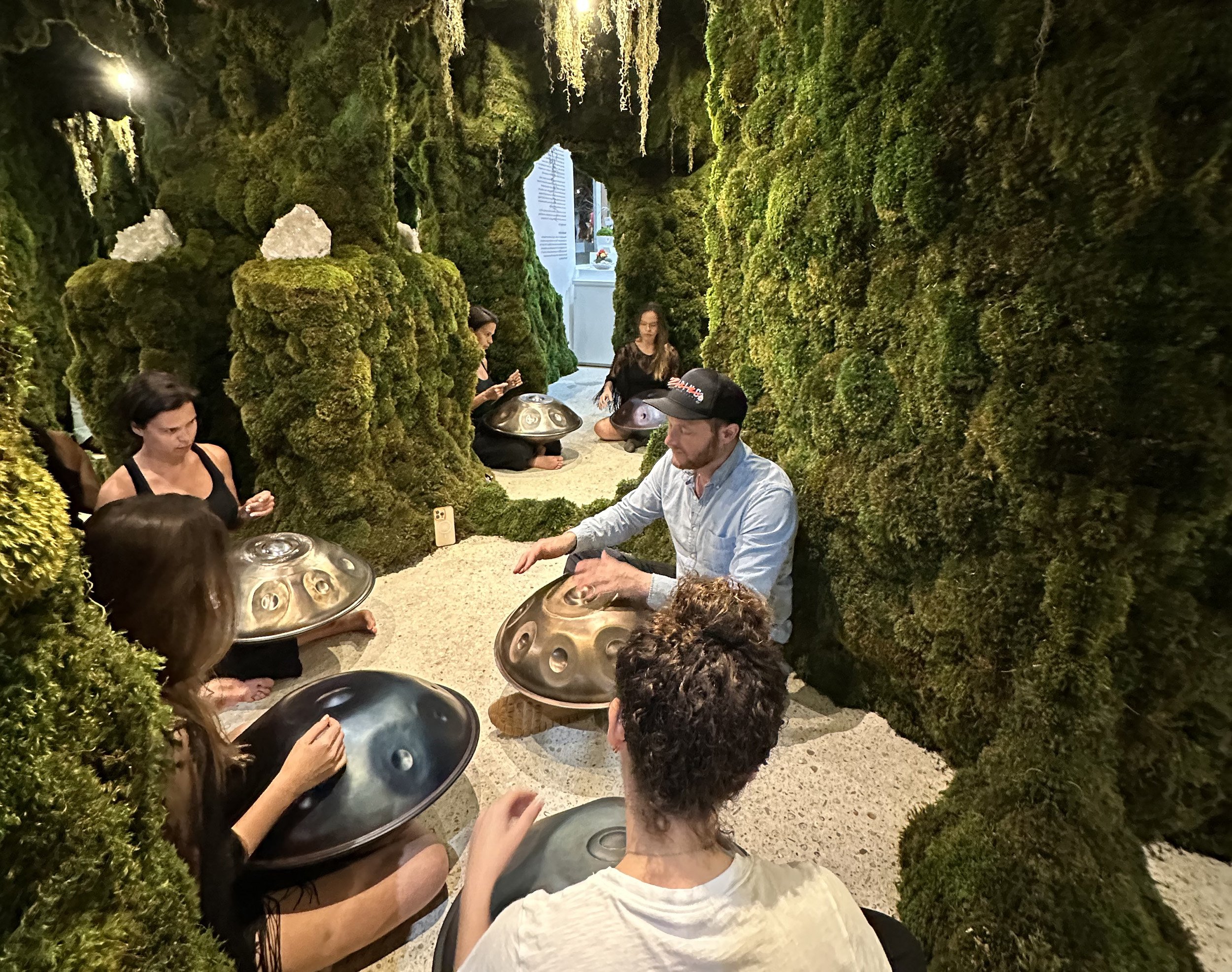 A group of people sitting in a room with green moss-covered walls, playing handpans. The setting resembles a natural, cave-like environment.