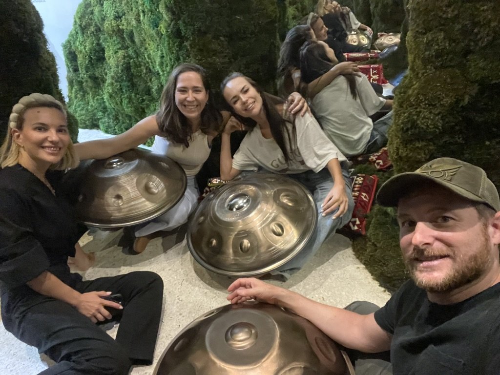 Group of four people sitting together holding handpans, surrounded by green, moss-like decor.