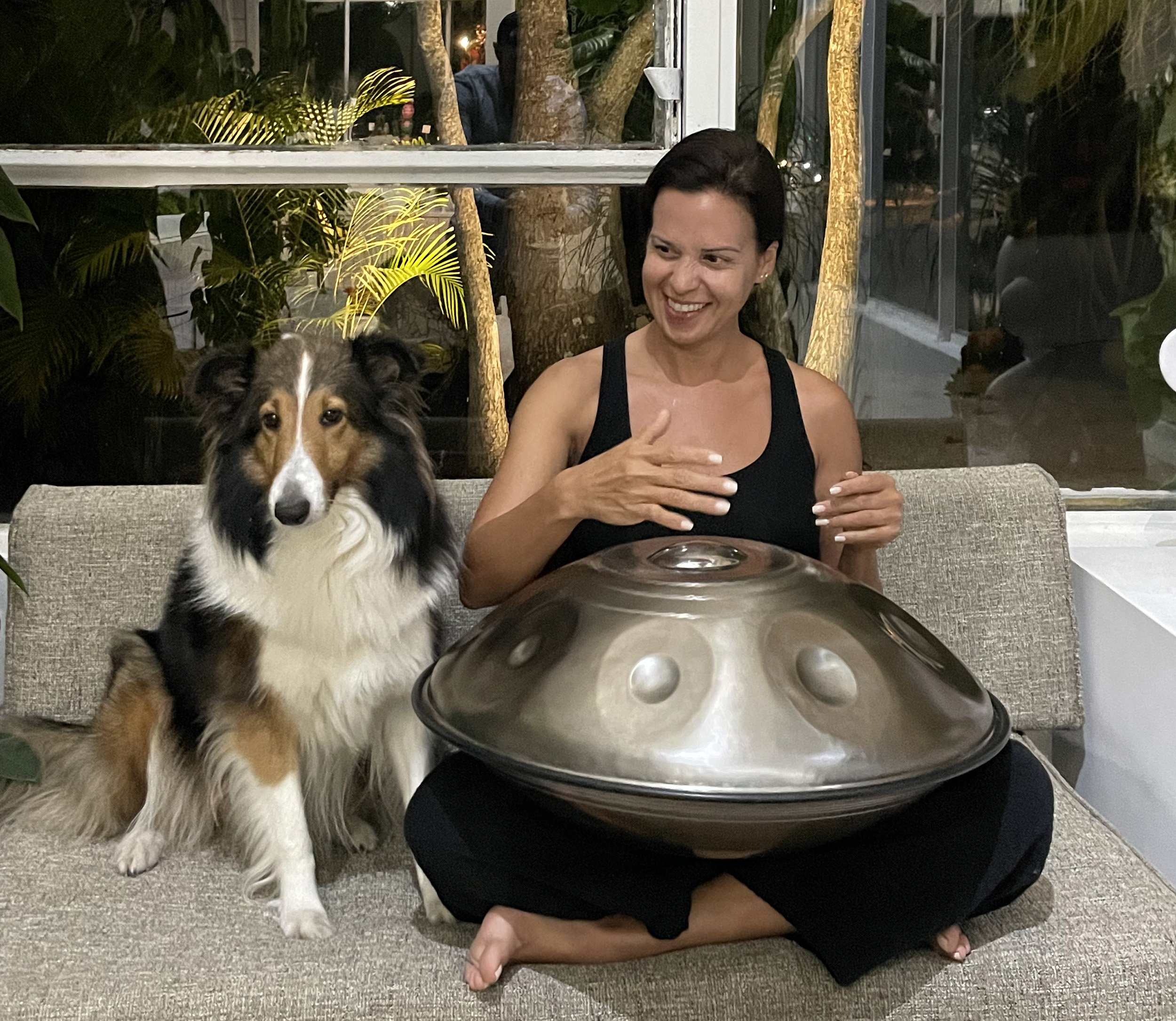 A woman sits on a sofa playing a hang drum next to a dog, indoors. The setting is cozy with plants and a window in the background.