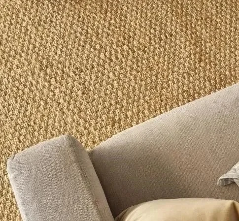Close-up view of a beige sofa with a striped pillow and a person's leg wearing beige pants resting on it, on a textured tan carpet.