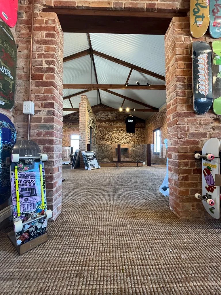 Skateboards mounted on brick walls and standing on the carpeted floor inside a spacious room with exposed beams and brick walls.