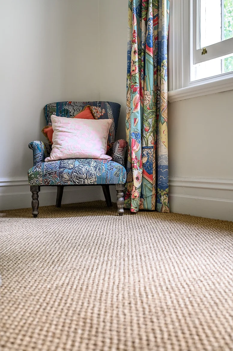 A patterned armchair with a pink pillow and an orange pillow beside a window with colorful curtains.