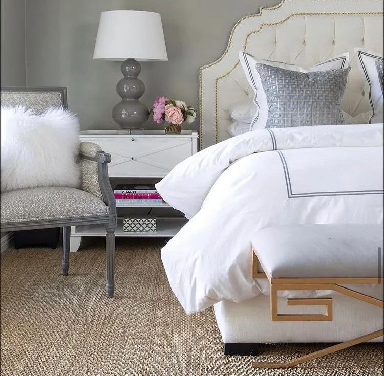 A bedroom corner with a white upholstered bed, gray decorative pillows, a white nightstand with pink and white flowers, a gray ceramic table lamp, a gray and white armchair with a white furry pillow, and a beige woven carpet.