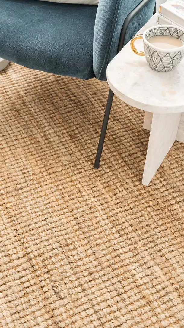 Close-up of a beige textured jute material installed on the floor, next to a small white marble side table with black and gold mug and some books, and part of a blue upholstered sofa with black metal leg.