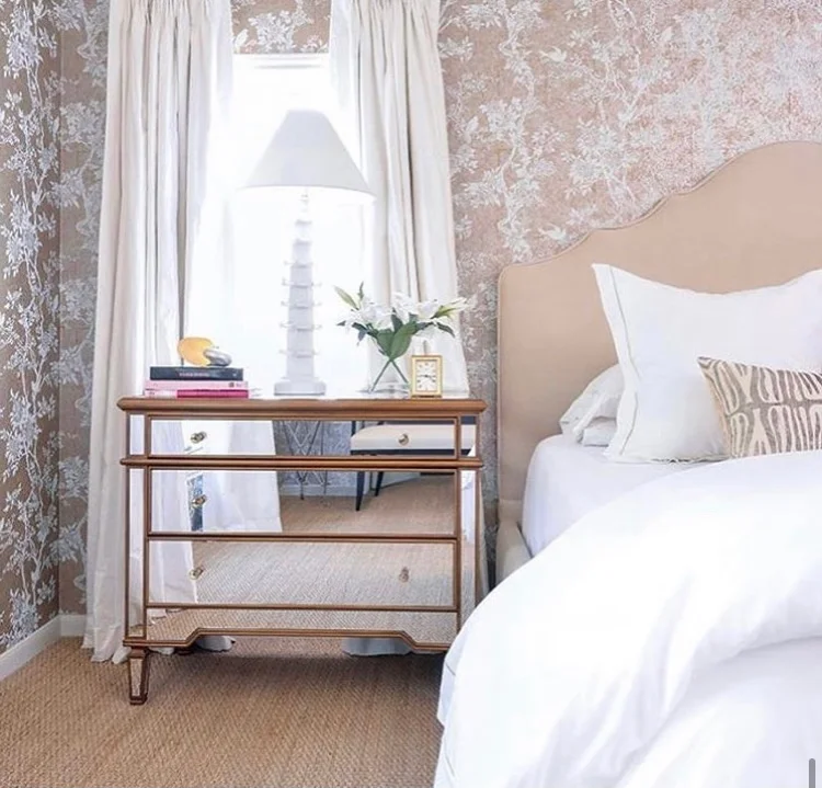 Bedroom corner with a beige bed, white pillows, a nightstand, a table lamp, a vase of white flowers, and decorative items, next to a window with curtains and floral wallpaper.
