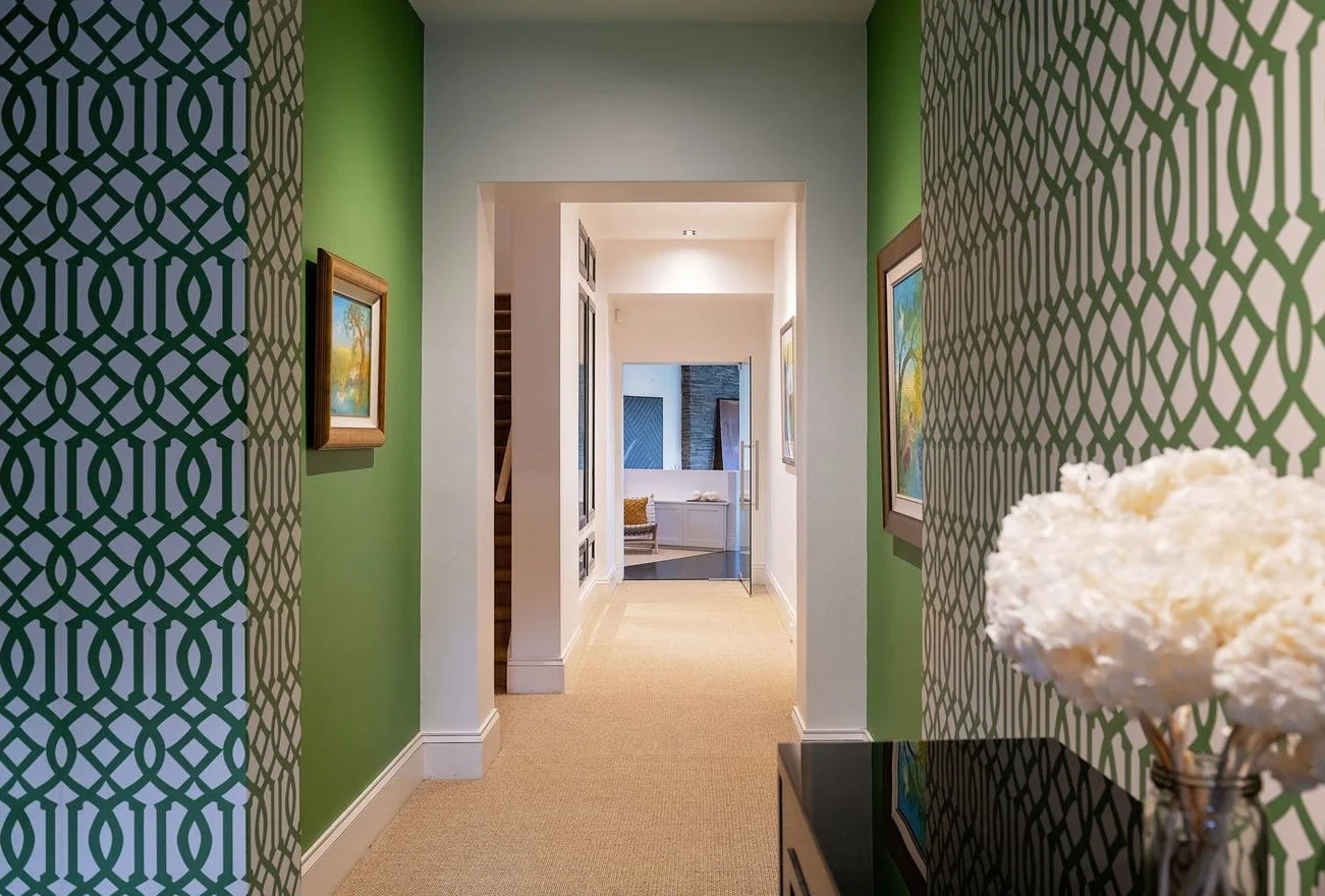 Hallway with green and patterned wallpaper walls, framed artwork, beige carpet, and a table with a vase of white flowers.