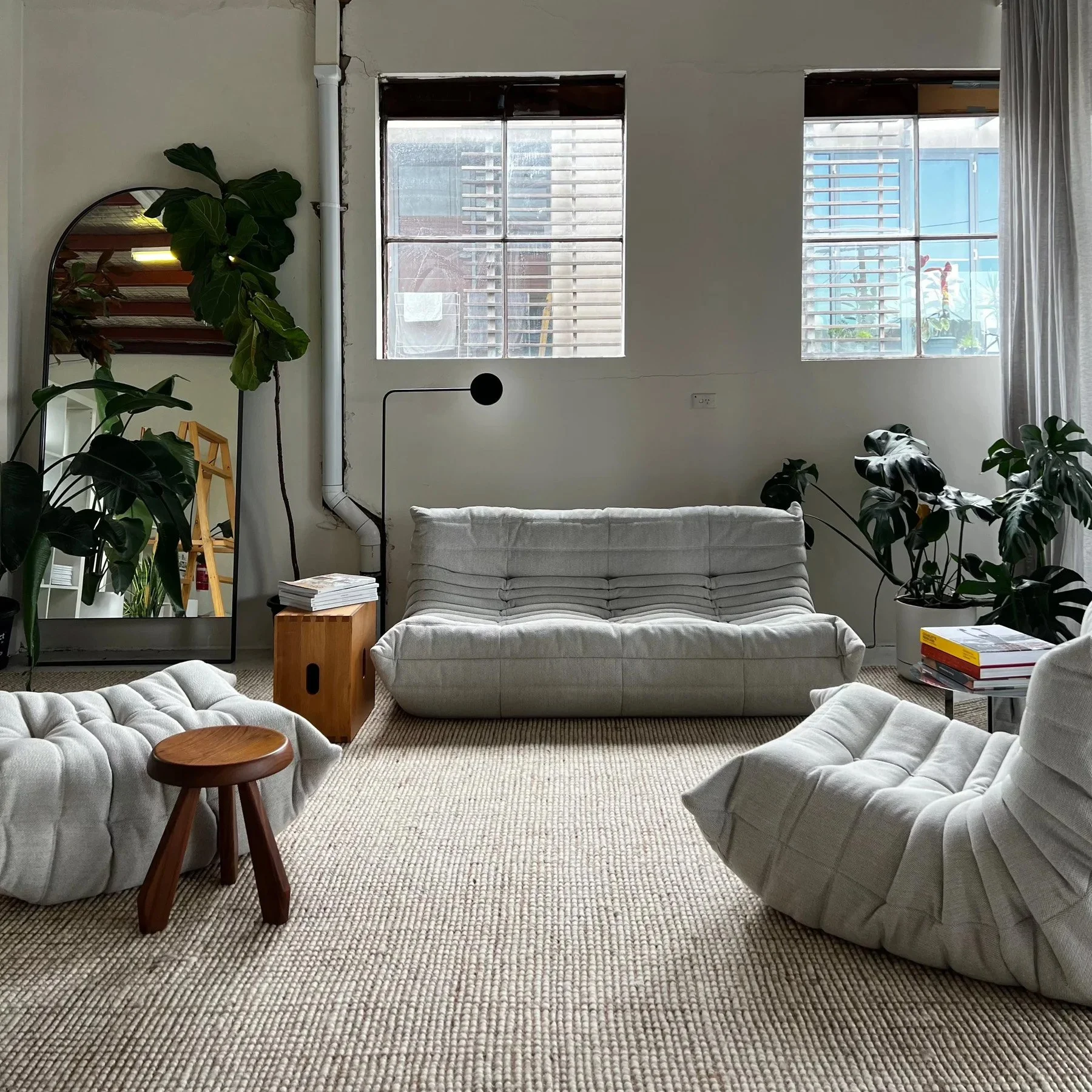 Living room with beige cushioned furniture, large plants, and two windows with blinds, sunlight coming through.
