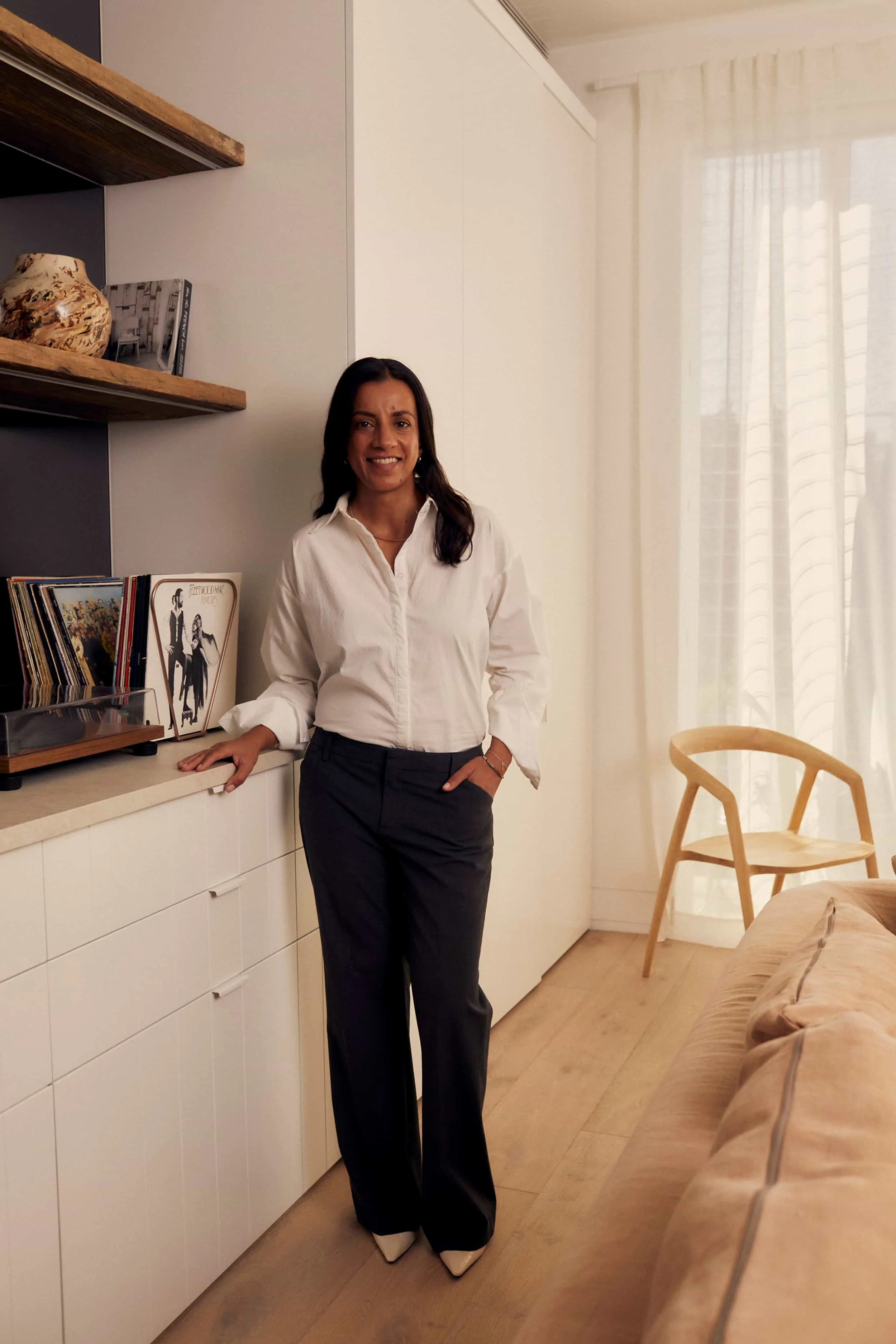 Dr Salina Jamani standing in a modern living room, smiling, wearing a white blouse, dark trousers, and white shoes, with vinyl records and album covers on a sideboard behind her.