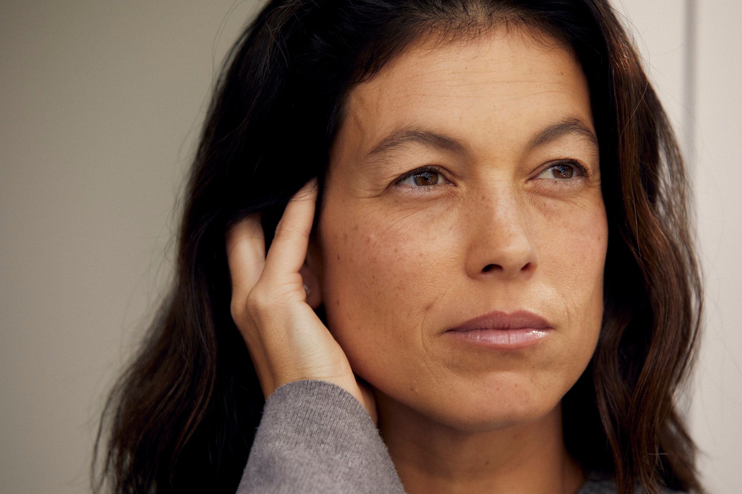 A woman with dark hair and light skin touches her ear with one hand, looking thoughtfully to the side.