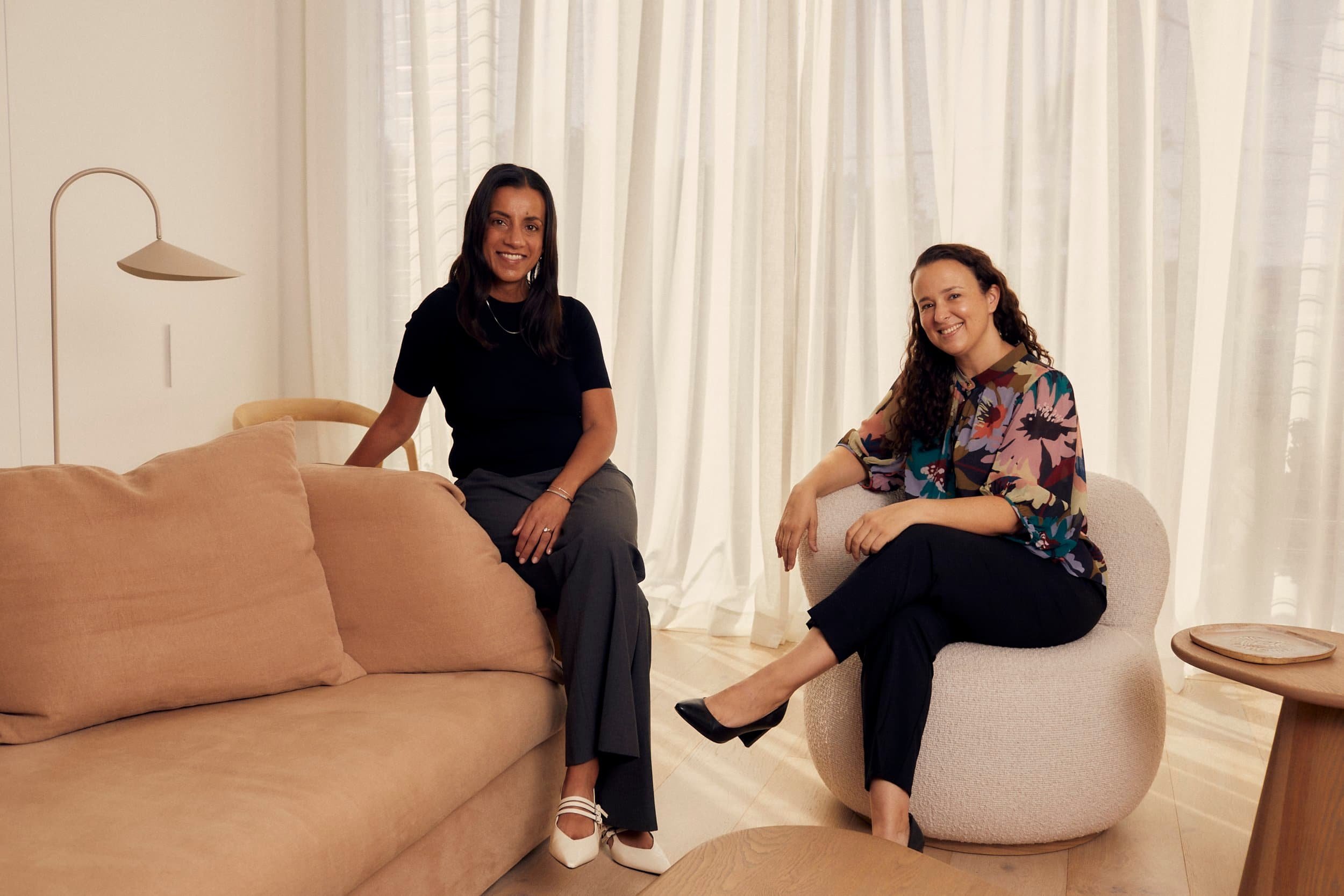 Dr Salina Jamani and Dr Caitlin Tunnicliffe sitting in a living room with beige and cream furnishings and white curtains, smiling at the camera.