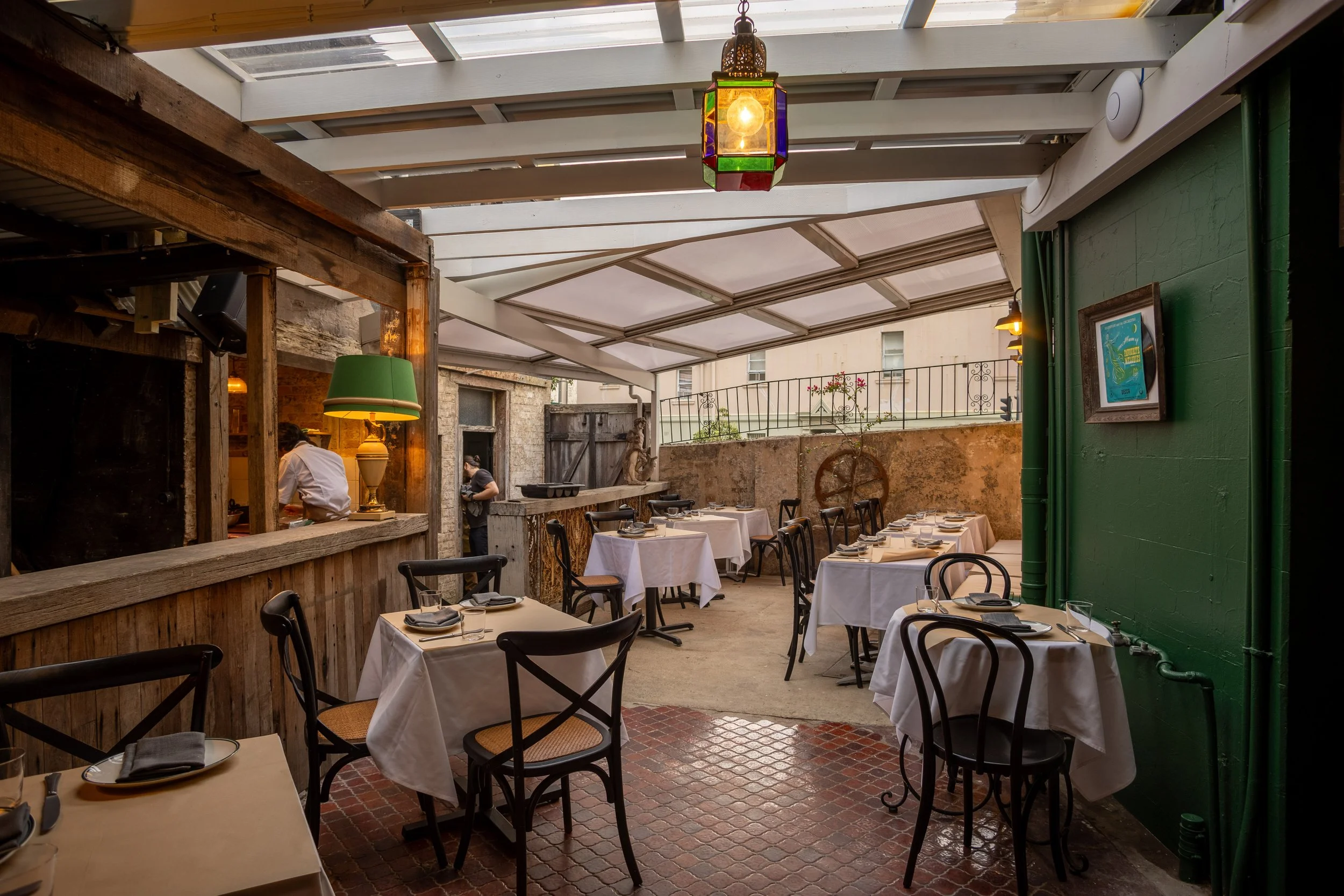Cozy restaurant interior with rustic decor, featuring wooden beams and stone walls, set up with tables covered with white tablecloths and black chairs. The space is well-lit, showing a mix of natural and artificial lighting. A chef is visible in the