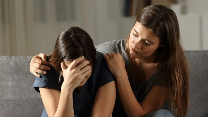 A woman console a young girl who is upset and crying, with her hand covering her face, in a home setting.