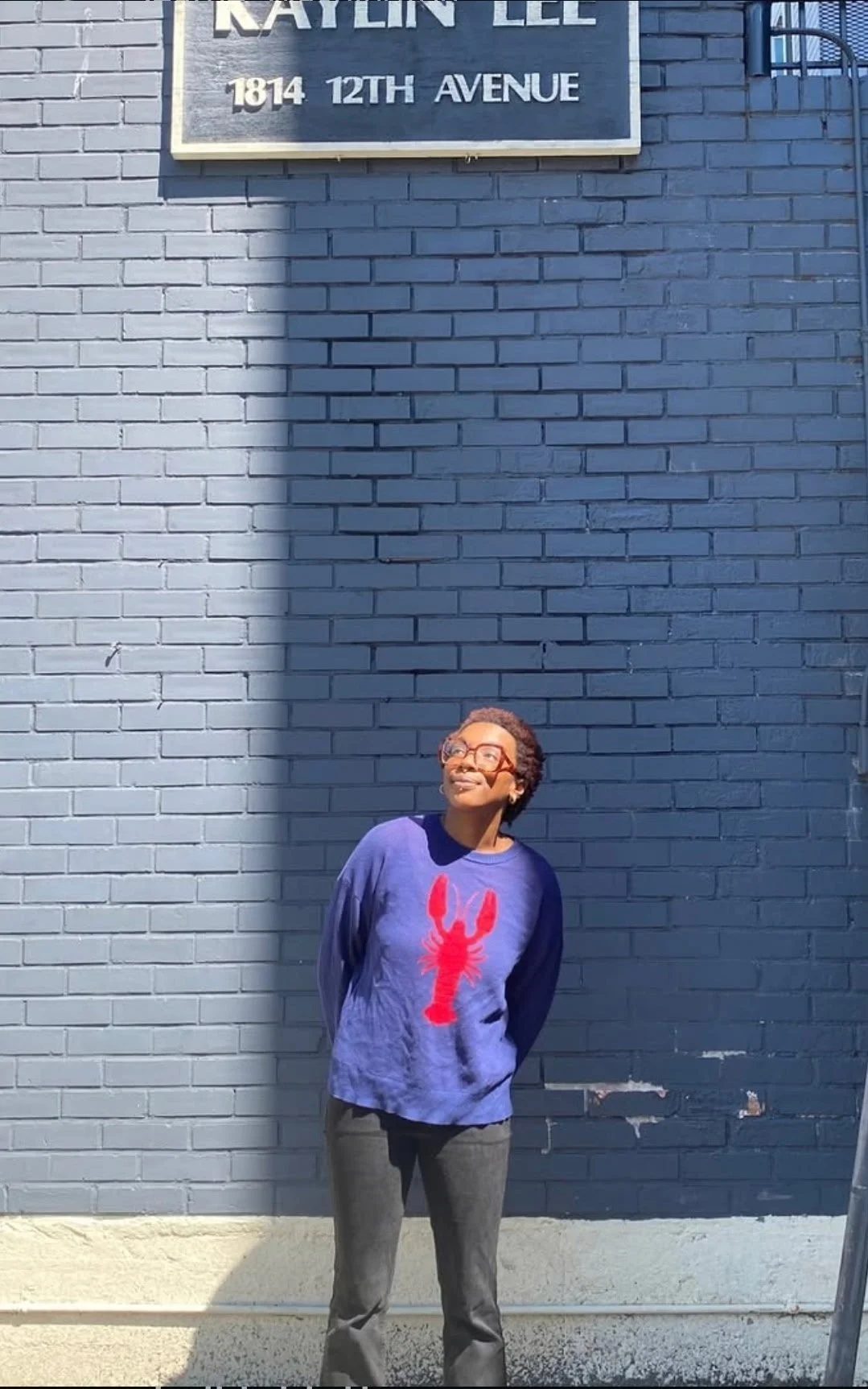 A woman with glasses and short hair standing against a blue brick wall, wearing a purple sweatshirt with a red lobster graphic on it, looking up with a pleasant expression.