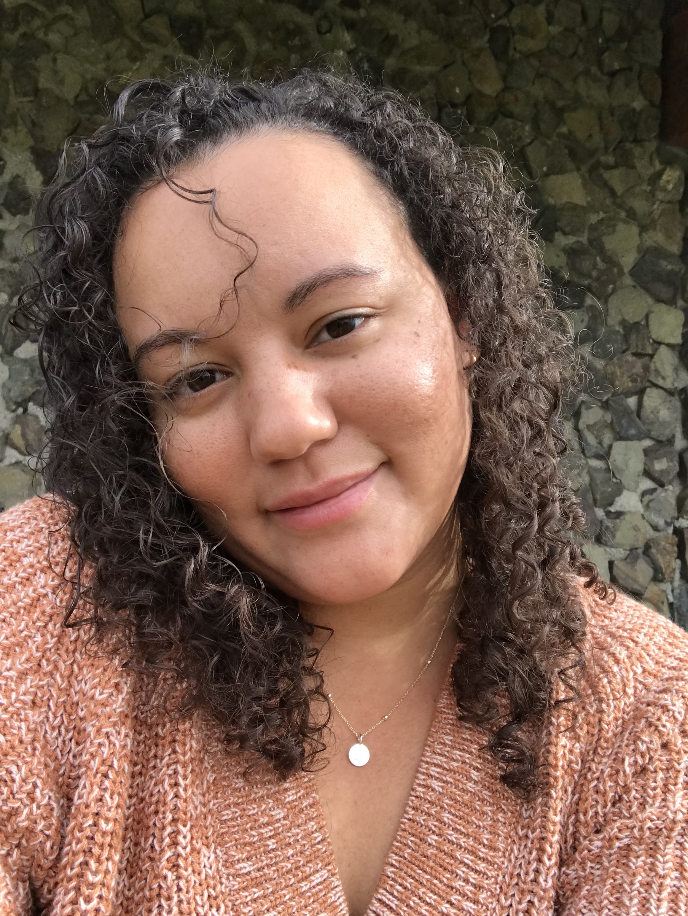 Close-up of a woman's face, with curly hair and wearing a peach cardigan and a necklace with a round pendant, with a stone wall in the background.