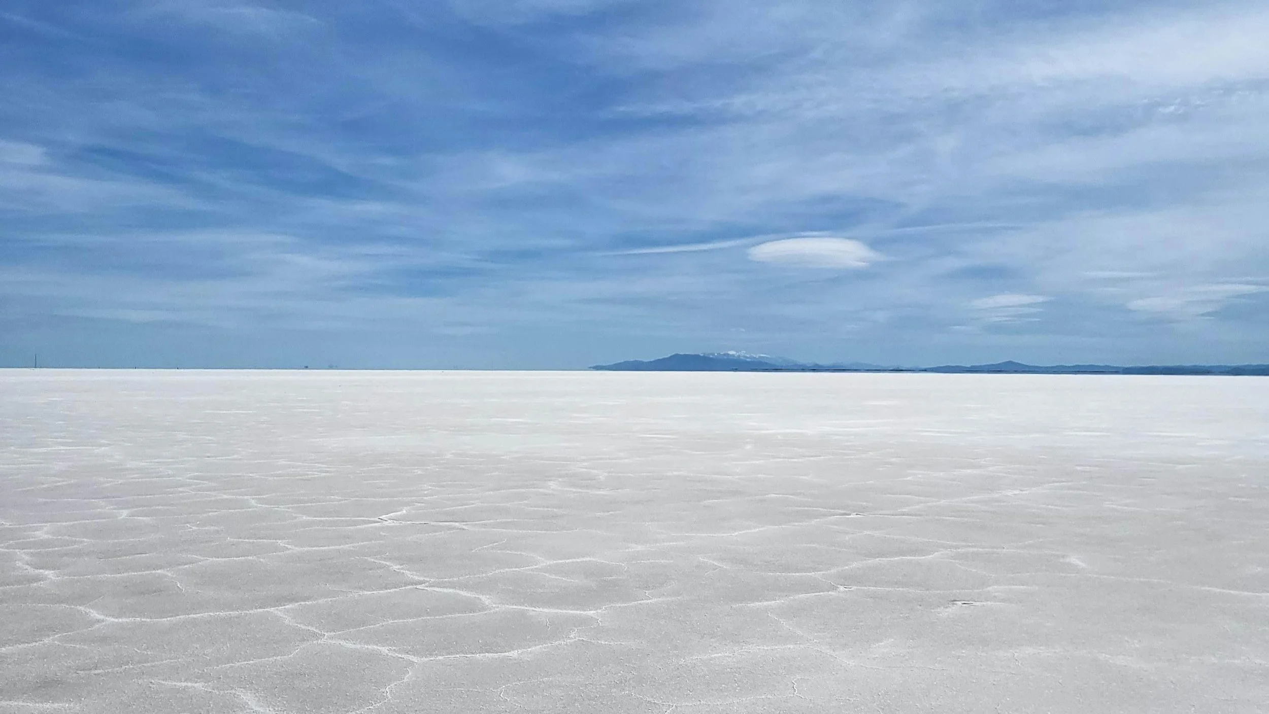 A vast, flat salt flat with white, cracked salt crust extending to the horizon, under a partly cloudy sky with distant mountains on the horizon.