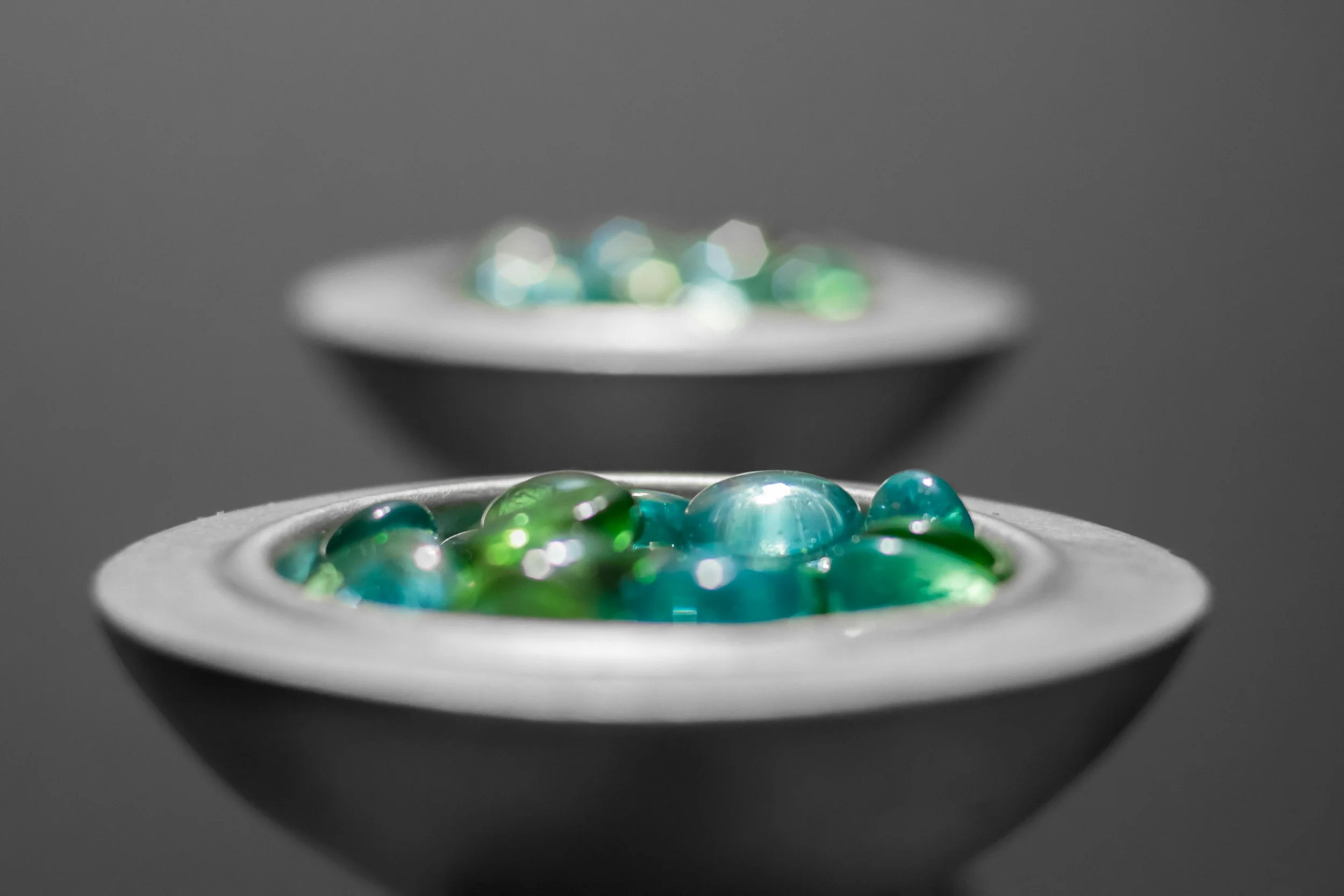 Close-up of colorful glass marbles in two small black bowls, with the front bowl sharply in focus and the background bowl slightly blurred, on a gray surface.