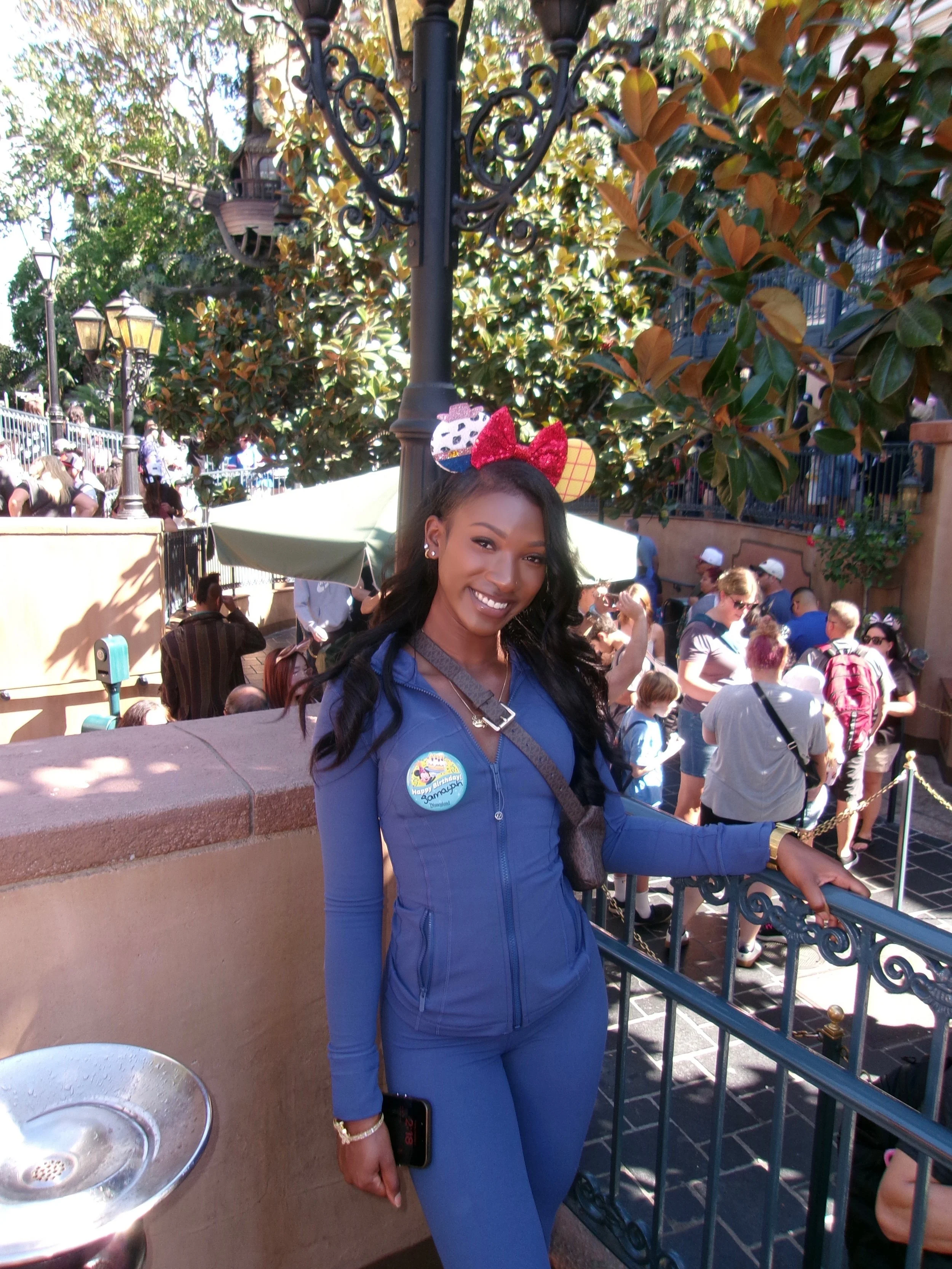 Young woman at Disneyland wearing a blue zip-up jacket and matching pants, with a Minnie Mouse ear headband, standing near a railing and smiling.