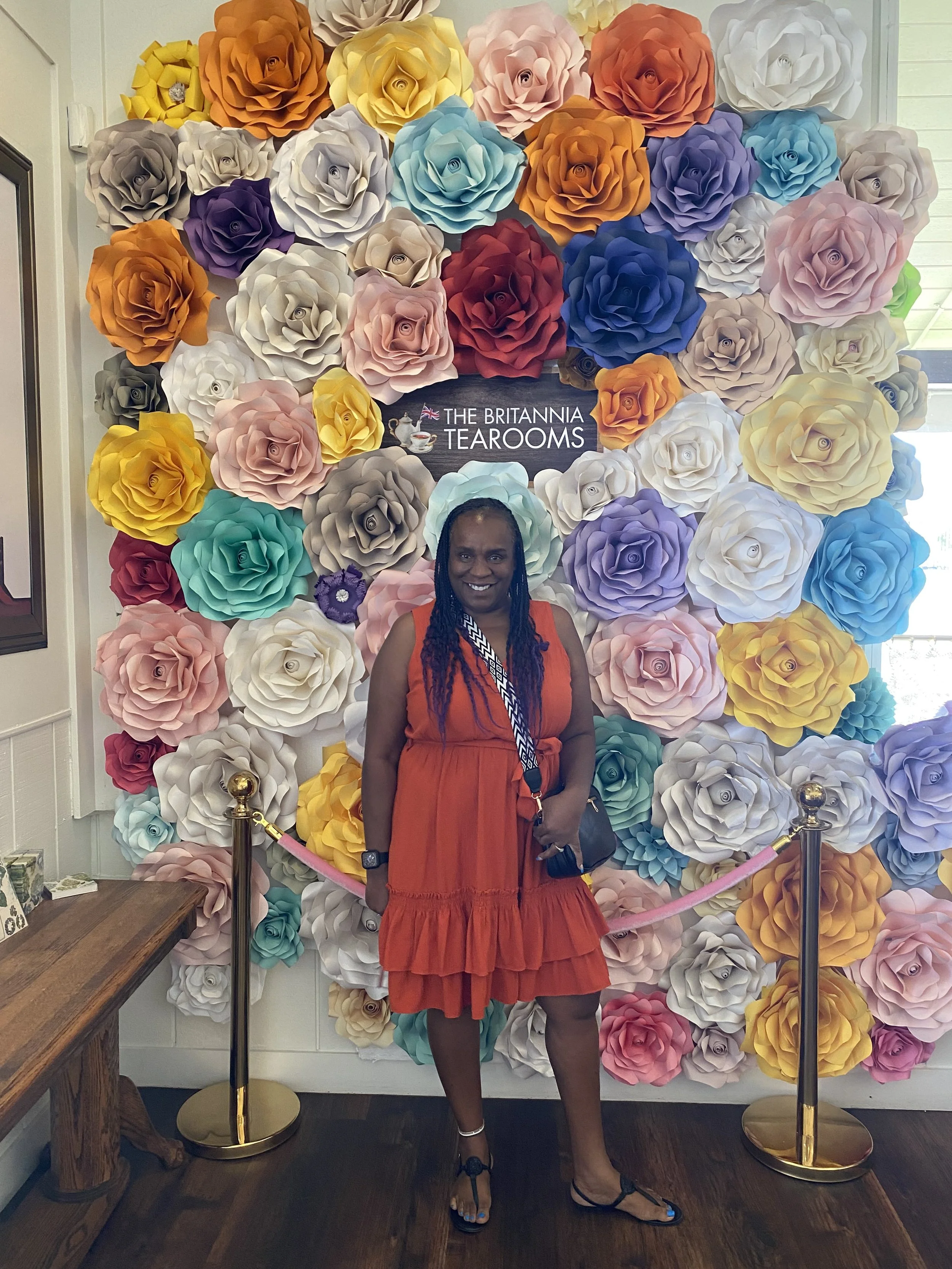 Woman standing in front of a wall covered with large, colorful paper roses at The Britannia Tearooms, smiling and wearing an orange dress with a lanyard and purse.