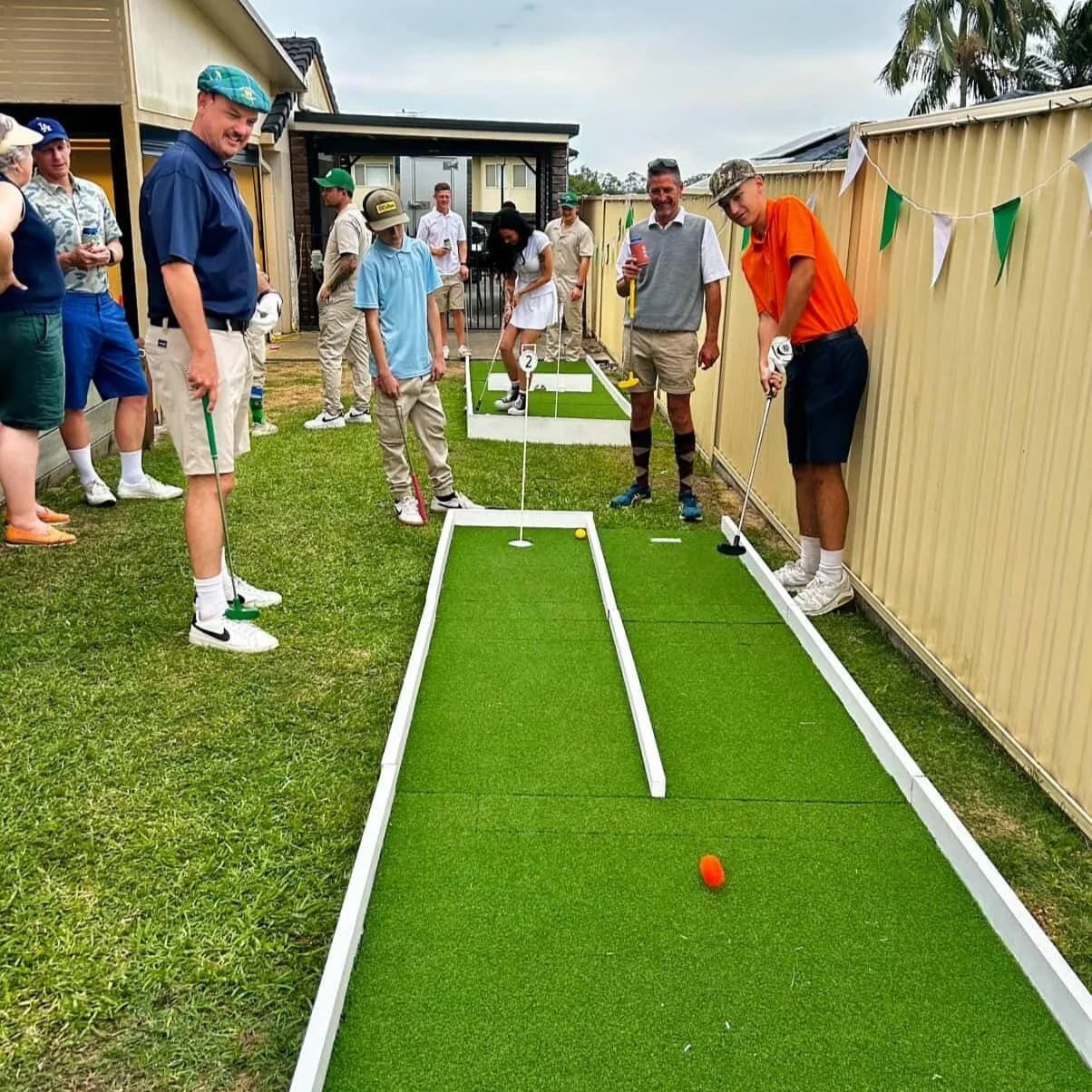 Group of people playing mini golf outdoors on a green artificial turf course, with some spectators watching.