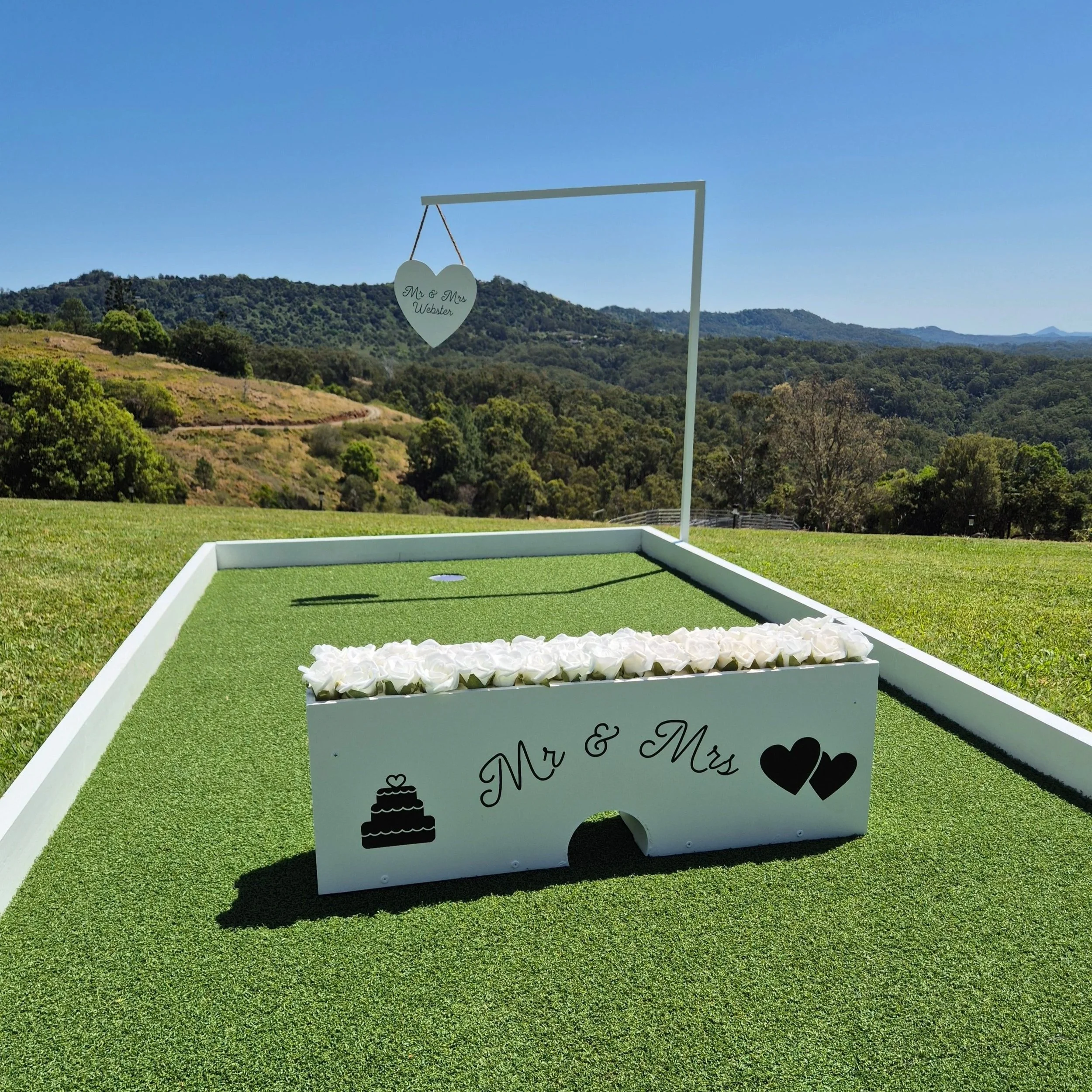 Wedding game setup with a white box labeled 'Mr & Mrs' and decorated with hearts and a wedding cake icon, with a hanging heart sign reading 'Mr & Mrs Webster', outdoors on a sunny day with rolling hills in the background.