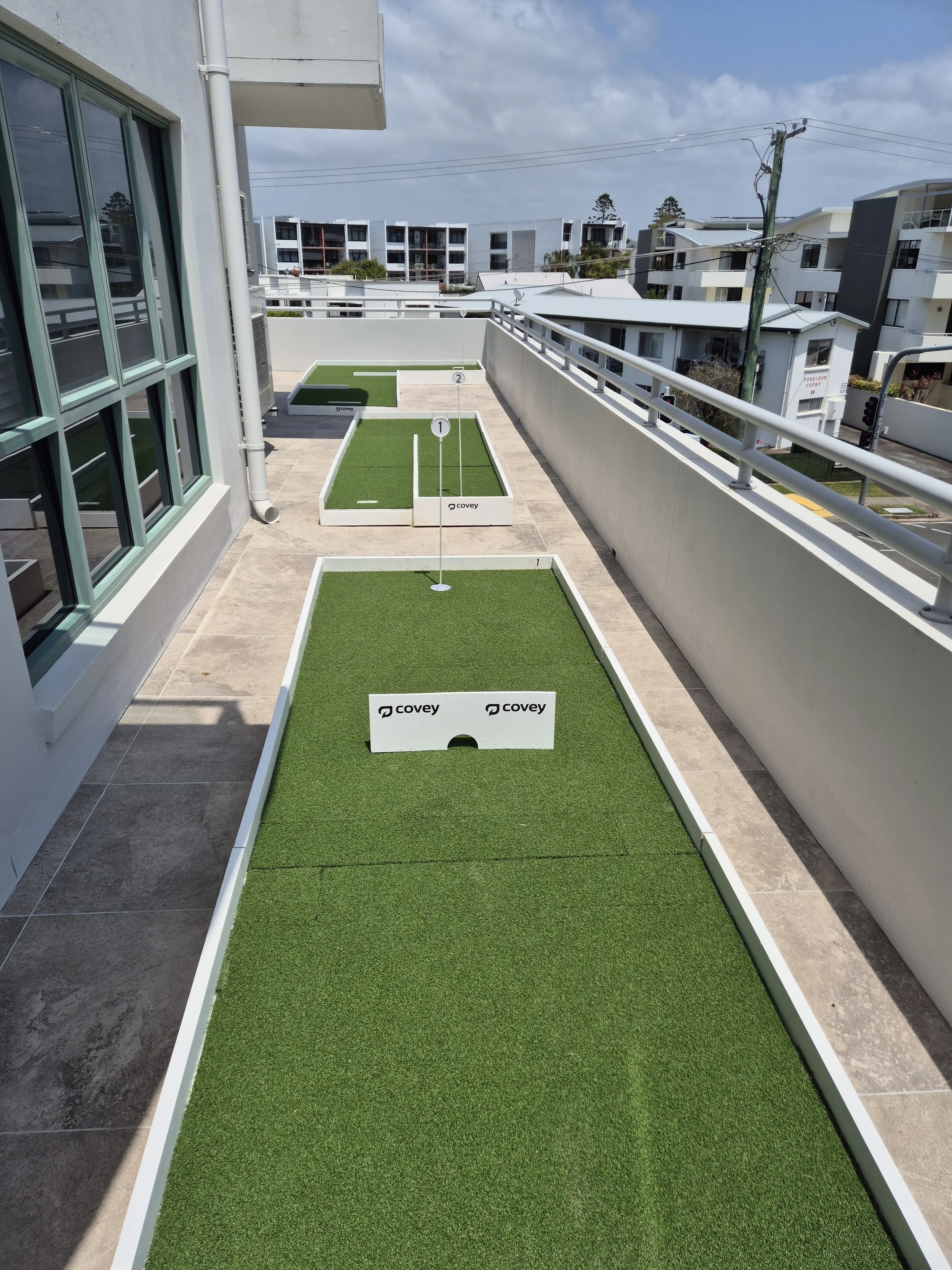 A rooftop mini golf course with three holes, each with artificial grass, white borders, and numbered signs, overlooking a cityscape of modern apartment buildings and power lines.