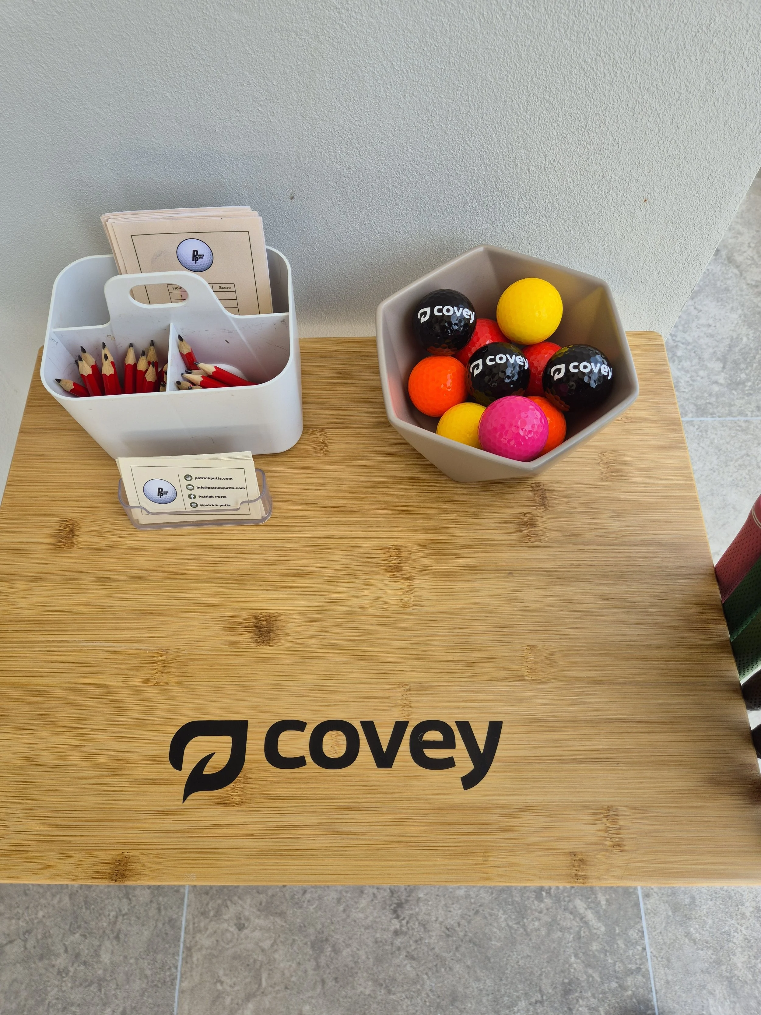A wooden table with the logo 'covey' printed on it, holding a white container with red and black pencils and a stack of papers, a small business card holder, and a gray bowl filled with colorful golf balls.