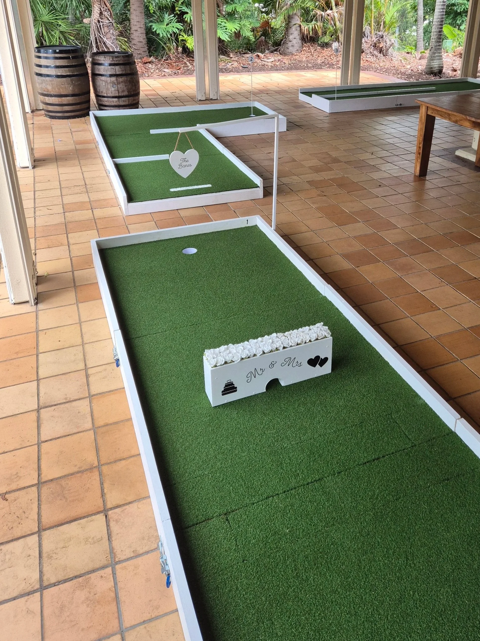 Miniature golf setup with three holes on a covered patio, surrounded by tropical plants and trees.