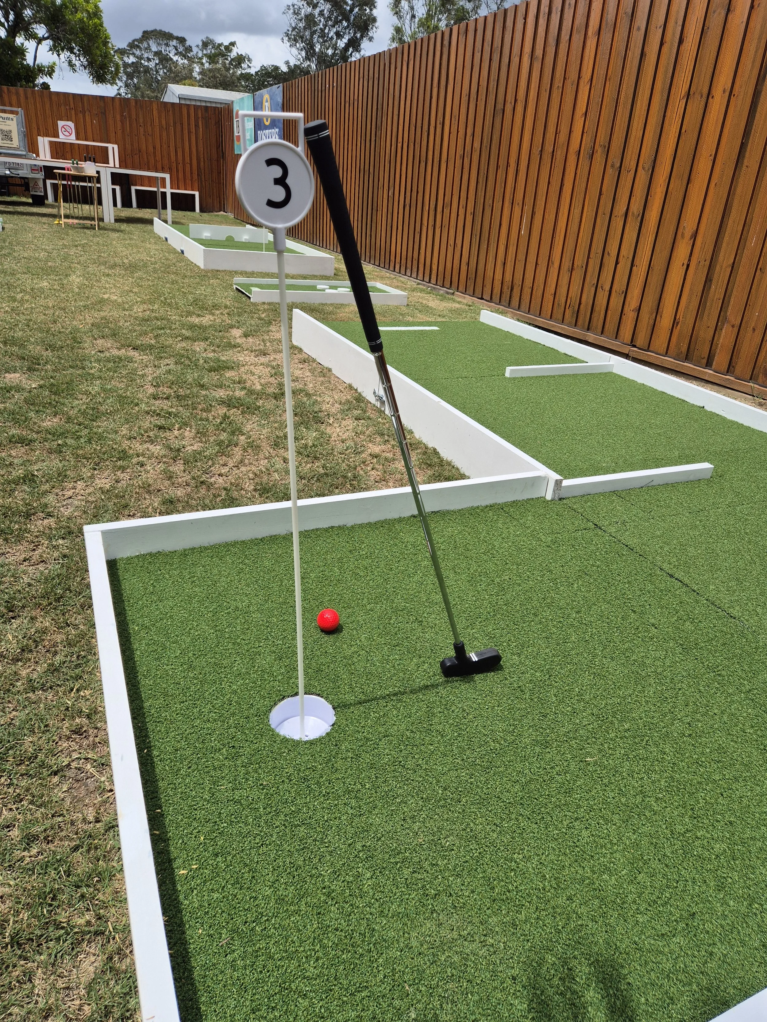 Mini golf putting green with a red golf ball, a flag with the number 3, and a golf club resting against the flagpole, outdoors with a wooden fence in the background.
