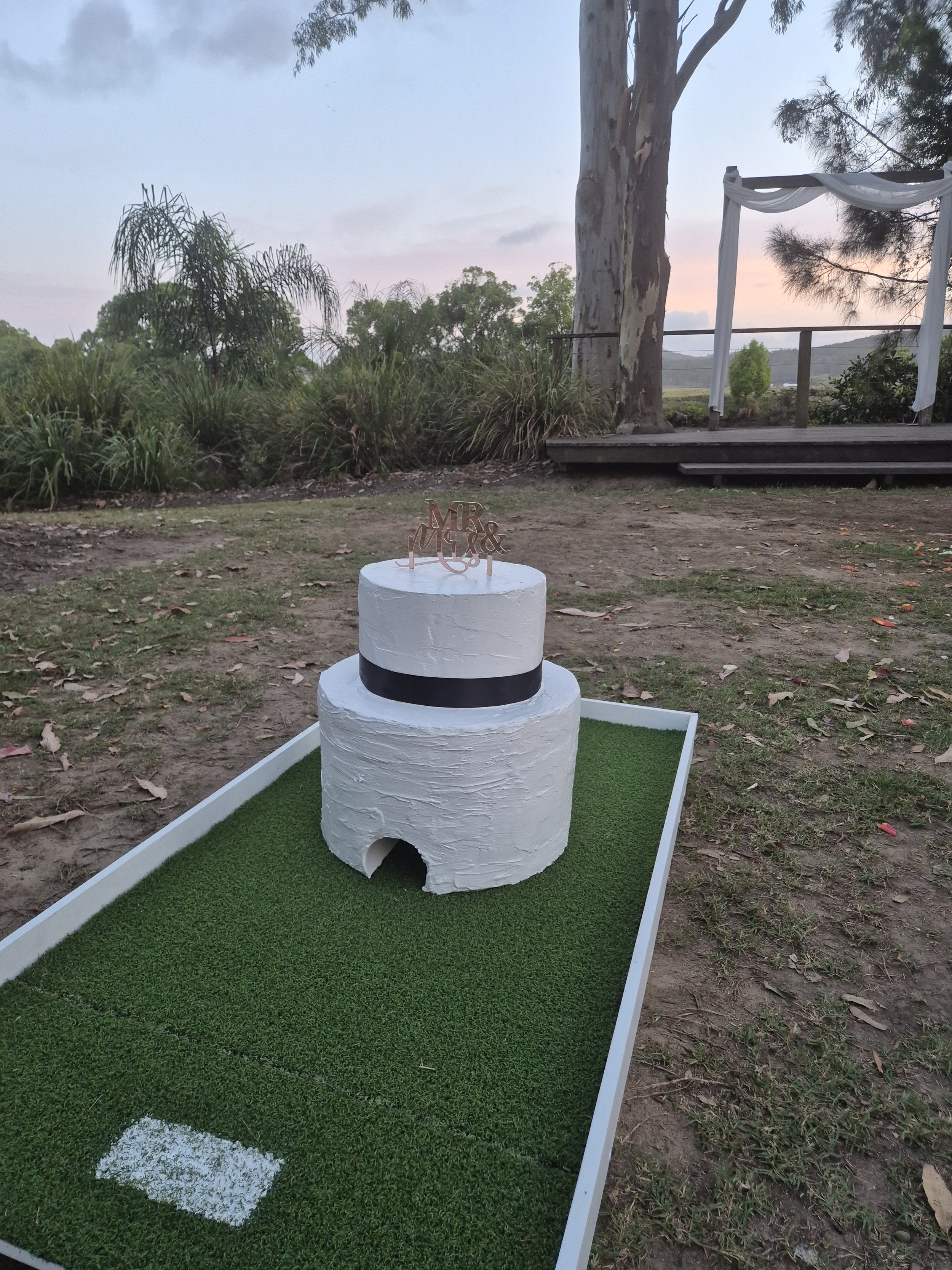 Miniature golf course with a small white cake decorated with a black band and a topper that reads "MR & MRS" placed on green artificial turf, outdoors with trees and a sunset sky in the background.