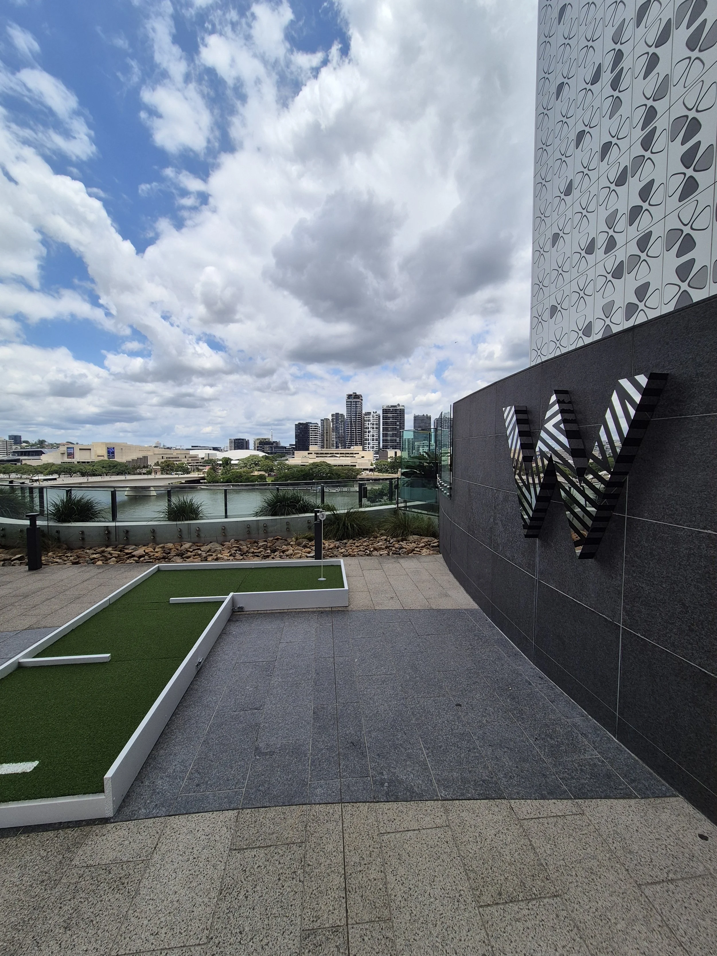 Urban rooftop garden with miniature golf course, city skyline with high-rise buildings, partly cloudy sky.