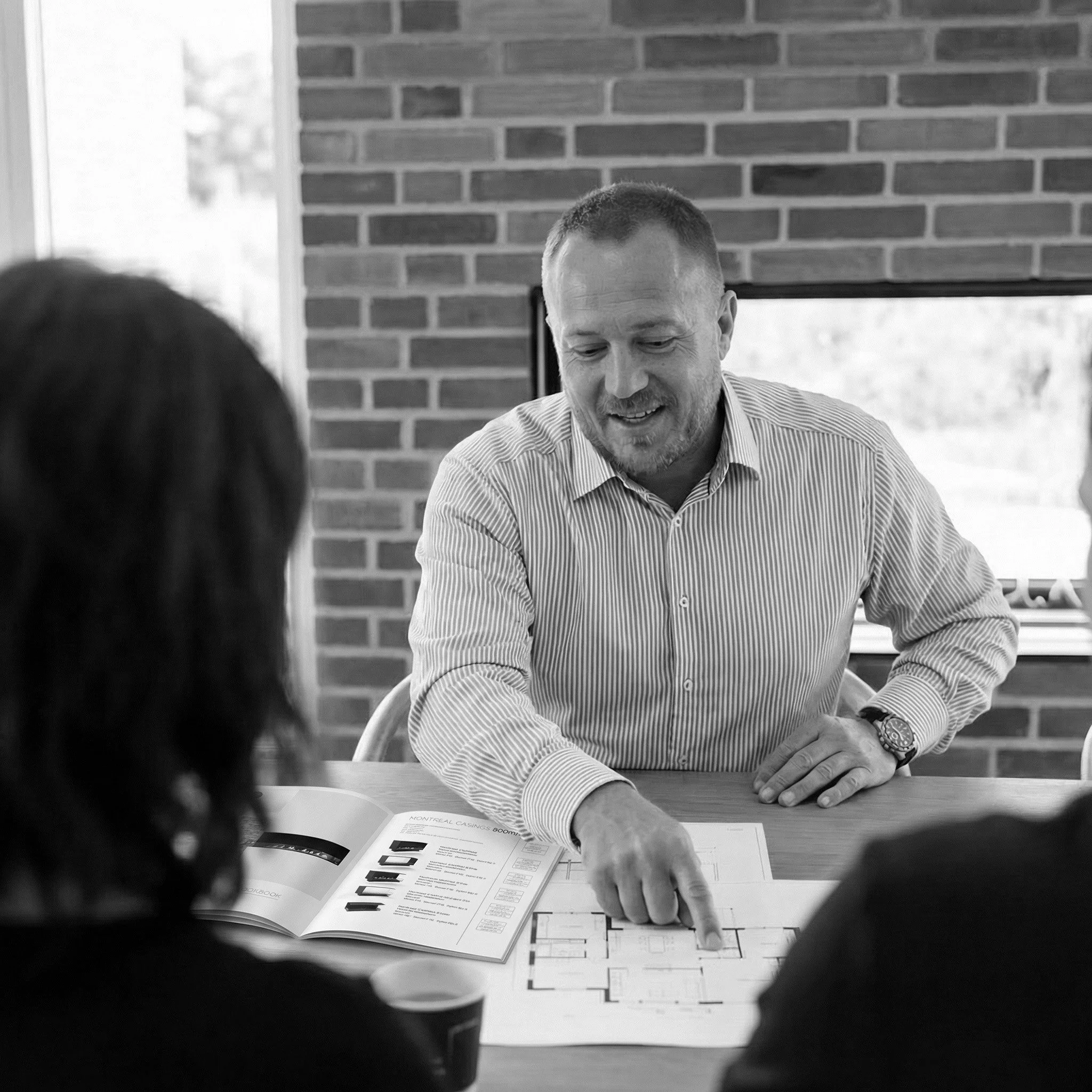 Black-and-white photo of a man in a shirt sitting at a table, pointing at a floor plan while speaking with two people seated opposite him. An open brochure and a coffee cup are on the table, and a brick wall forms the background.