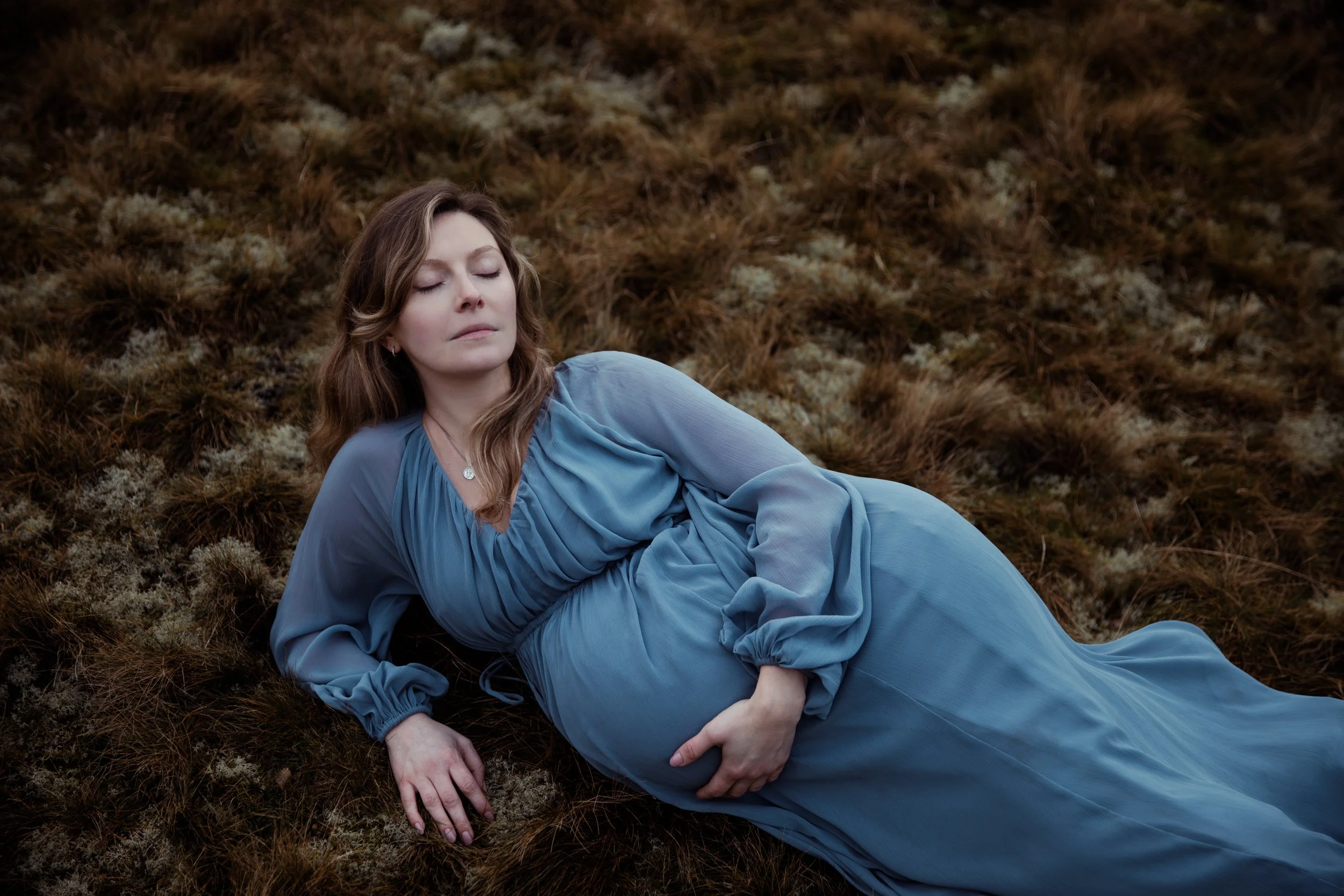 A woman with brown hair in a blue dress with long sleeves, lying on the ground covered with brown grass and moss, with her eyes closed and one hand on her stomach.